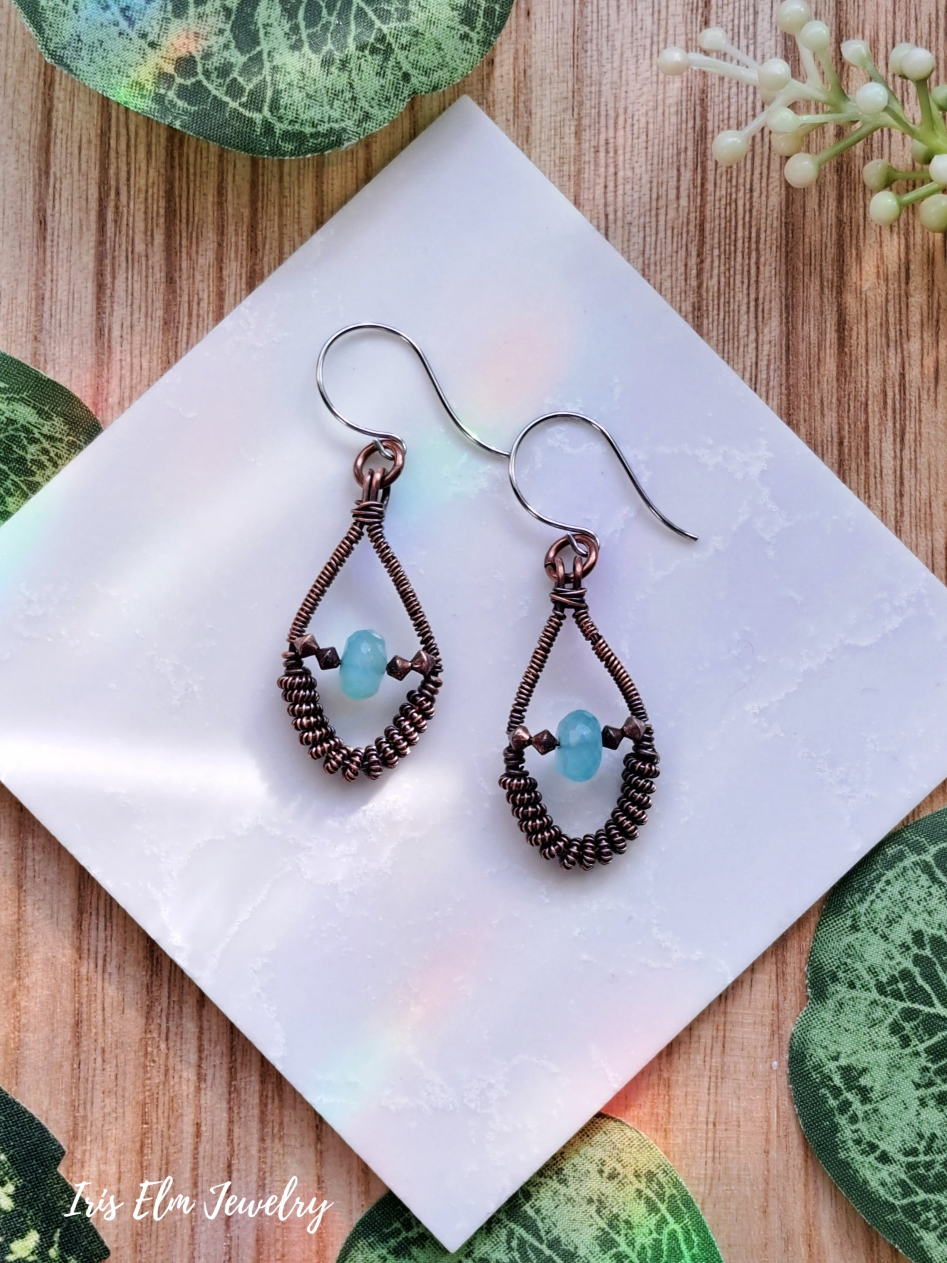 Pair of copper wire wrapped coil teardrop earrings with blue chalcedony beads on a white napkin with green leaves in the background