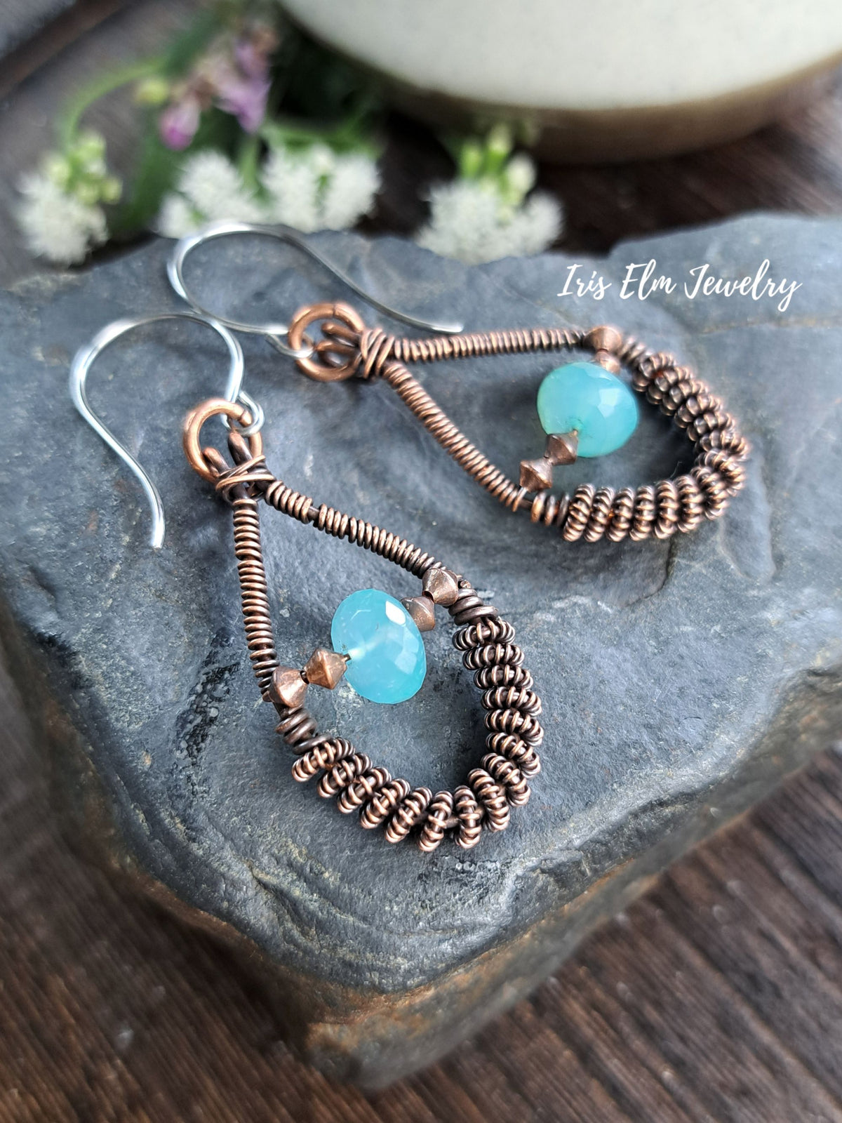 A pair of handmade dangle earrings by Iris Elm Jewelry, featuring copper wire wrapped teardrop shapes with light blue faceted beads and silver fishhook wires, displayed on a dark textured stone.