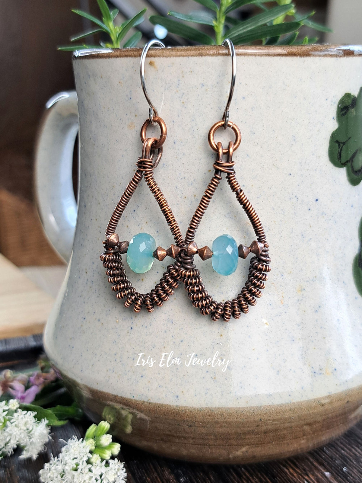 A pair of handmade copper wire dangle earrings with light blue faceted beads, displayed on a speckled ceramic mug with green foliage in the background.