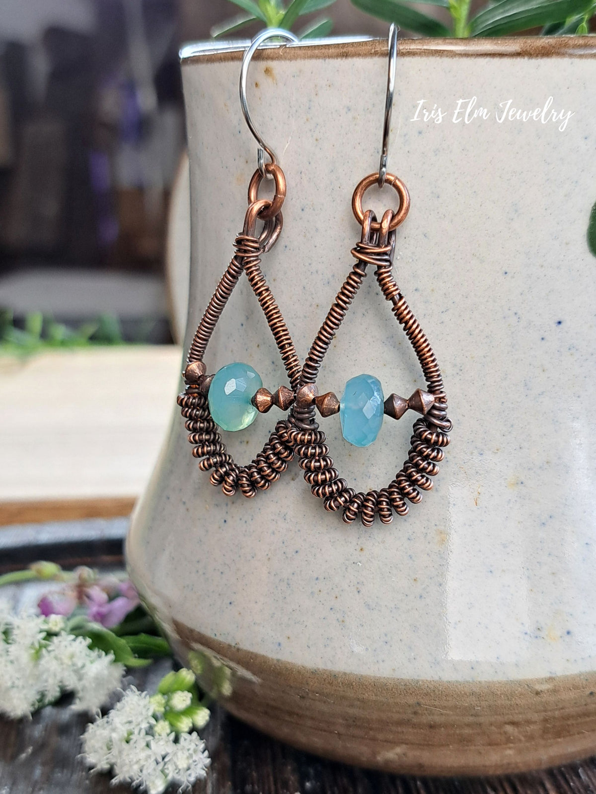 A pair of handmade copper wire teardrop dangle earrings with light blue faceted beads, hanging from silver fishhook wires. The earrings are displayed in front of a speckled ceramic pot with a blurred natural background.
