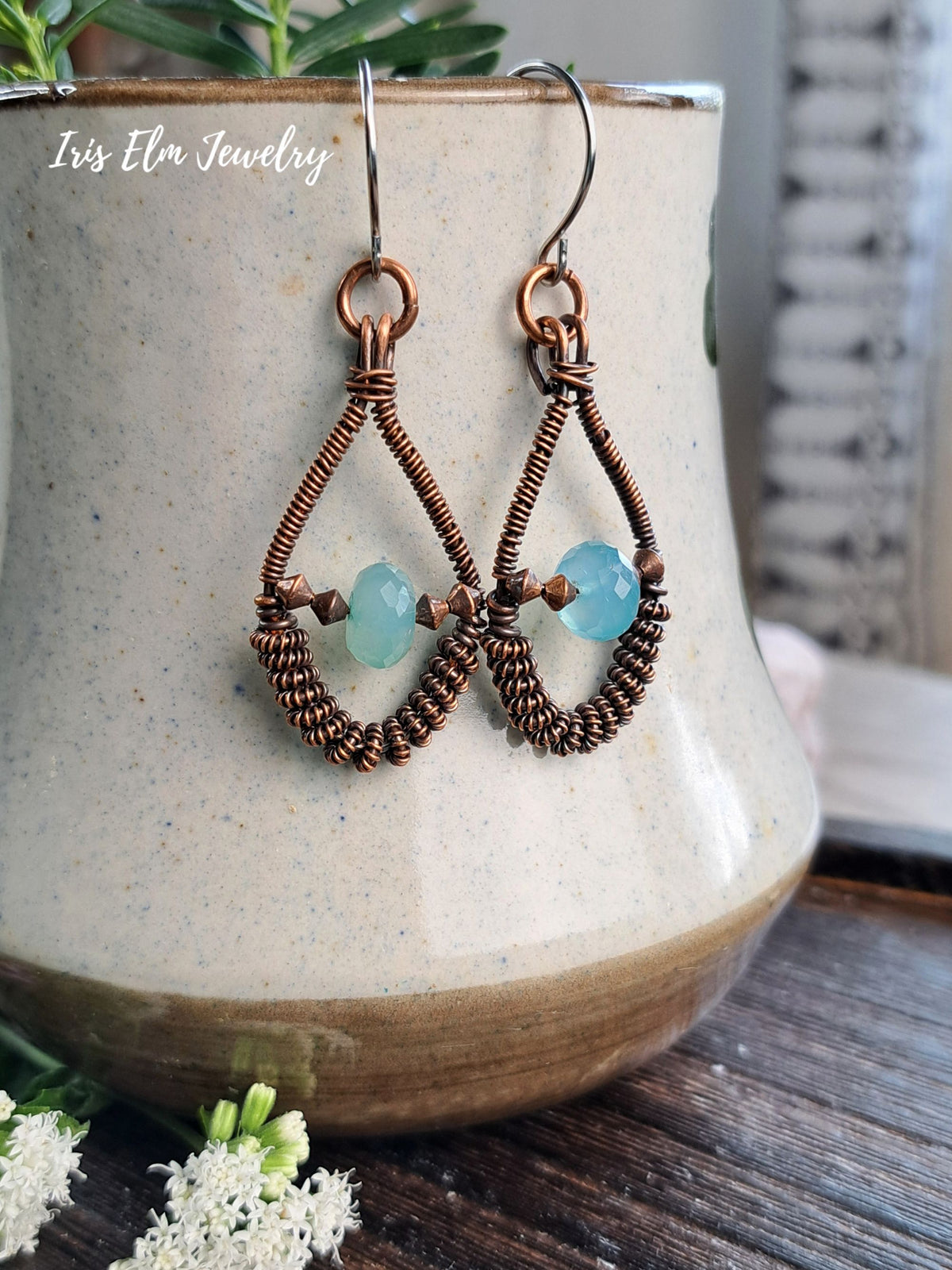 A pair of handmade copper wire dangle earrings with light blue faceted beads, hanging from silver fishhook wires. They are displayed draped over a beige ceramic pot on a dark wooden surface.