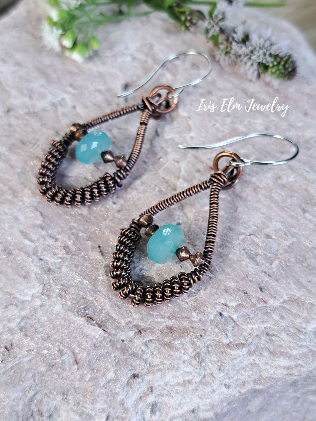 A pair of handmade teardrop dangle earrings with copper wire wrapping, small brown beads, and light blue-green stone accents, displayed on a textured stone surface with a white script watermark reading 'Iris Elm Jewelry'.
