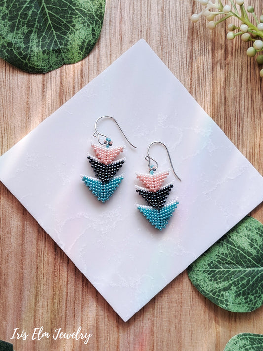 Pair of beaded 3 tier triangle seed bead pink, grey, and blue earrings on a white card with green leaves on a wooden surface