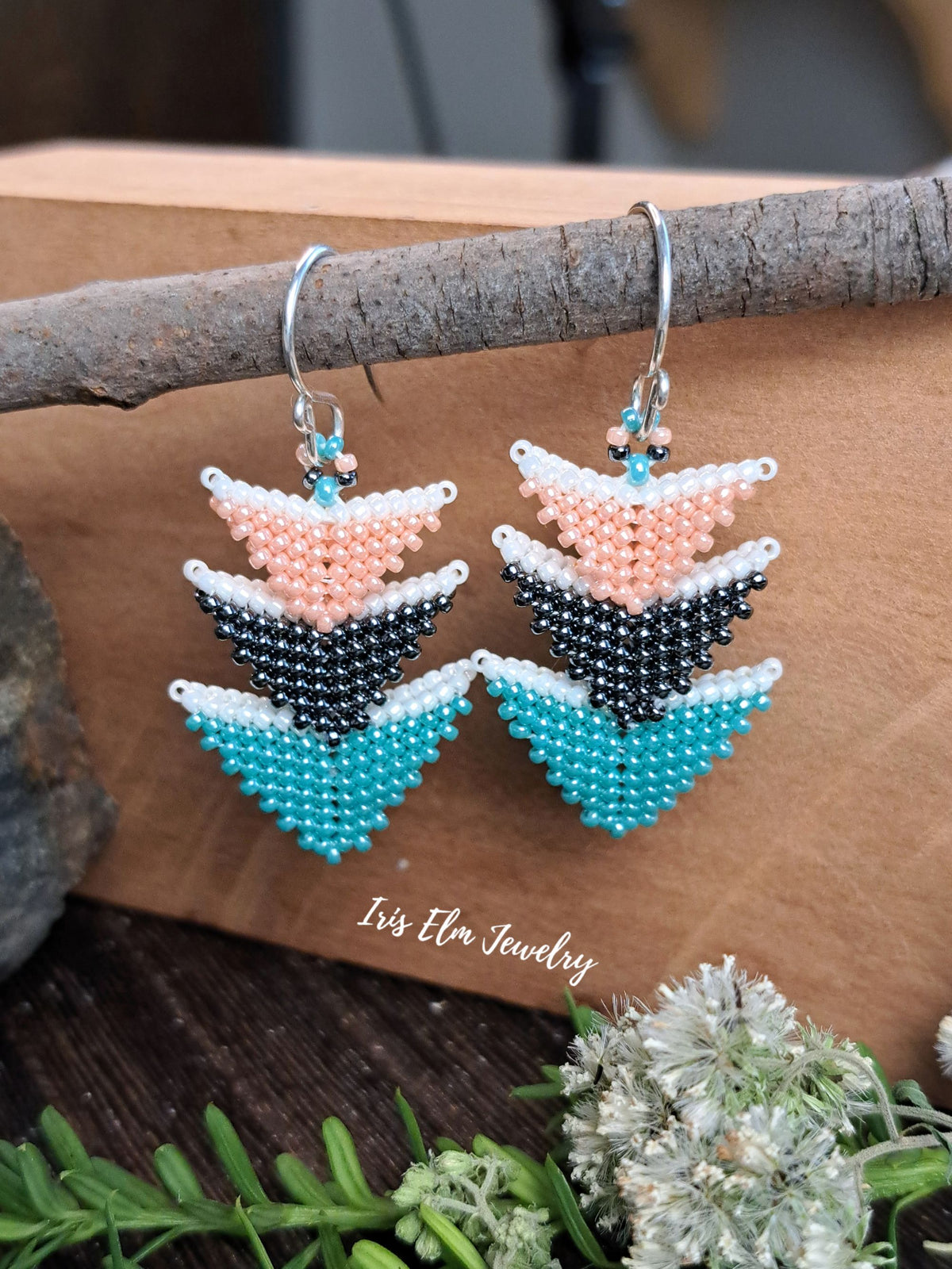 A pair of handmade beaded dangle earrings with a geometric chevron pattern in turquoise, black, peach, and white, hanging from silver hooks. The earrings are displayed on a rustic wooden branch with a brown background and green foliage at the bottom.