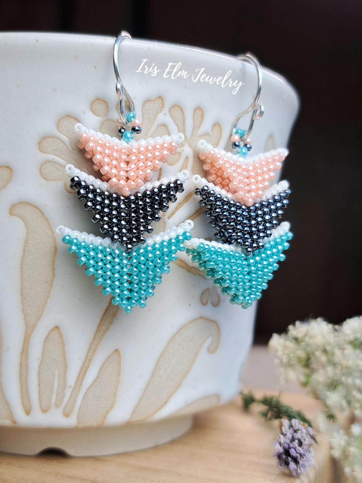 A pair of handmade beaded earrings by Iris Elm Jewelry, featuring three chevron layers in peach, black, and turquoise, displayed on a white ceramic pot.