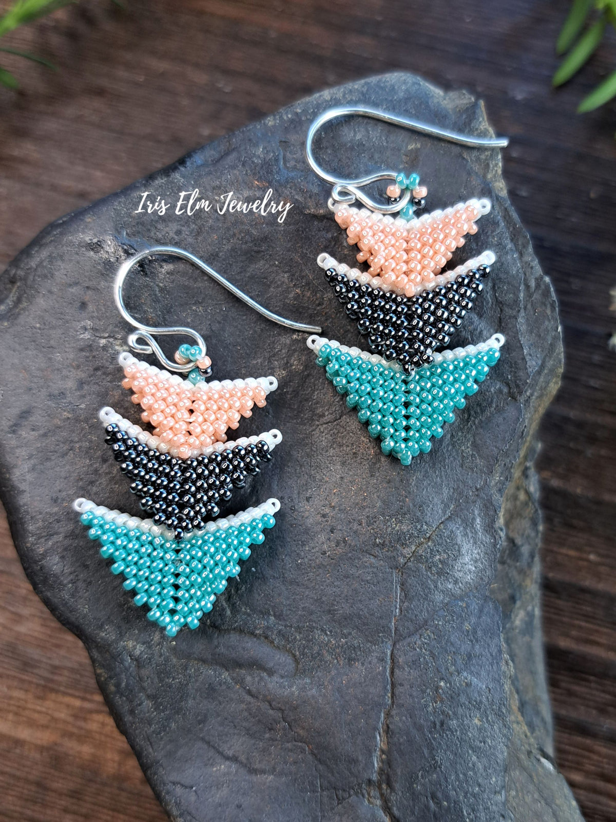A pair of handmade beaded dangle earrings with a geometric chevron pattern in peach, black, and turquoise, displayed on a dark textured stone. The earrings feature silver-tone fishhook wires 