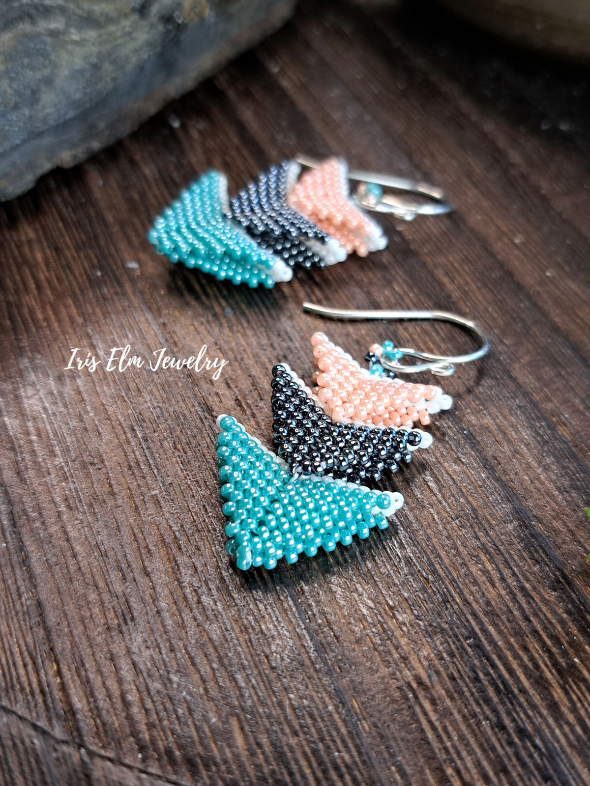 Three pairs of handmade beaded earrings with triangular chevron designs in teal, black-and-white, and peach, displayed on a dark wooden surface. The brand name 'Iris Elm Jewelry' is watermarked on the left.