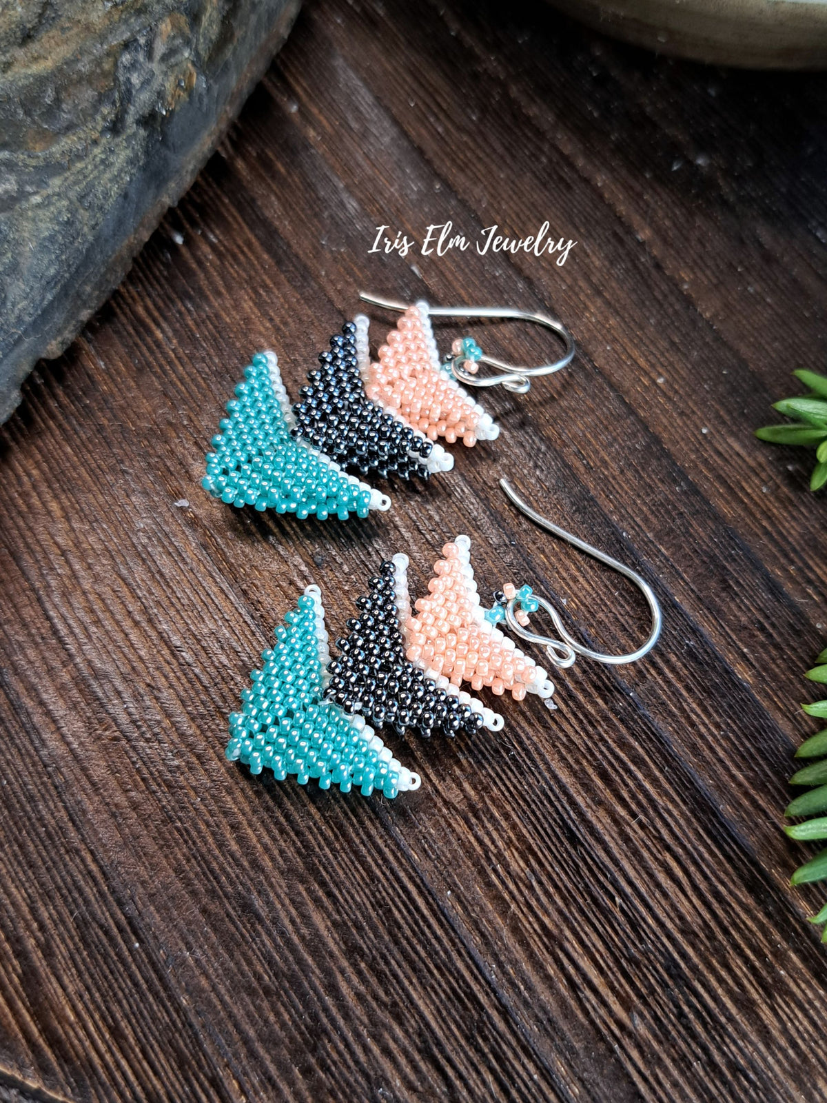 Pair of handmade triangle beaded dangle earrings with chevron patterns in turquoise, black, and peach, displayed on a dark wood surface with a brand watermark 'Iris Elm Jewelry'.