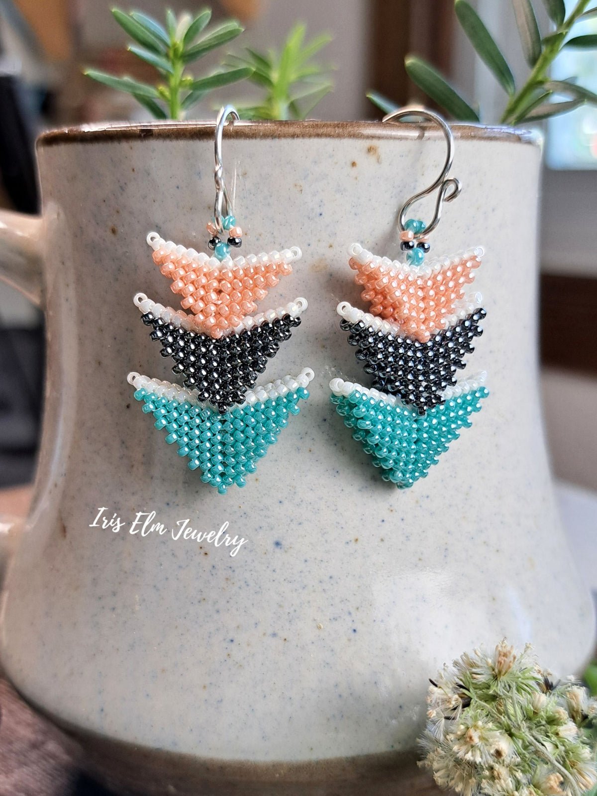 A pair of handmade beaded drop earrings with a stacked chevron design in peach, black, and teal, hanging from silver fishhook wires. They are displayed on the front of a speckled ceramic mug with green plants in the background.