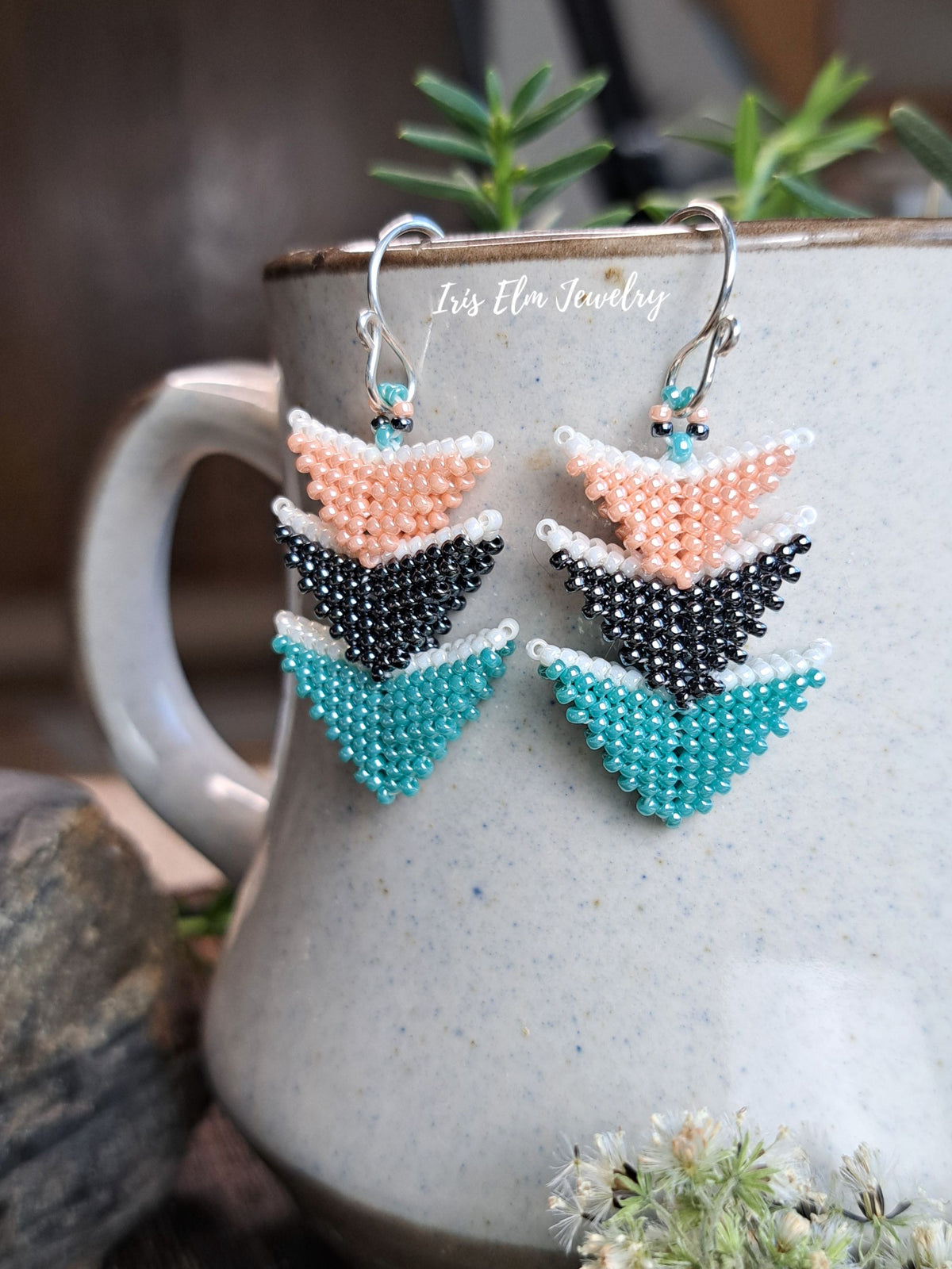 A pair of handmade beaded dangle earrings with a geometric chevron pattern in peach, grey, and teal, hanging from silver fishhook wires. The earrings are displayed on the side of a speckled gray ceramic mug with rosemary sprigs and a stone prop in the background.