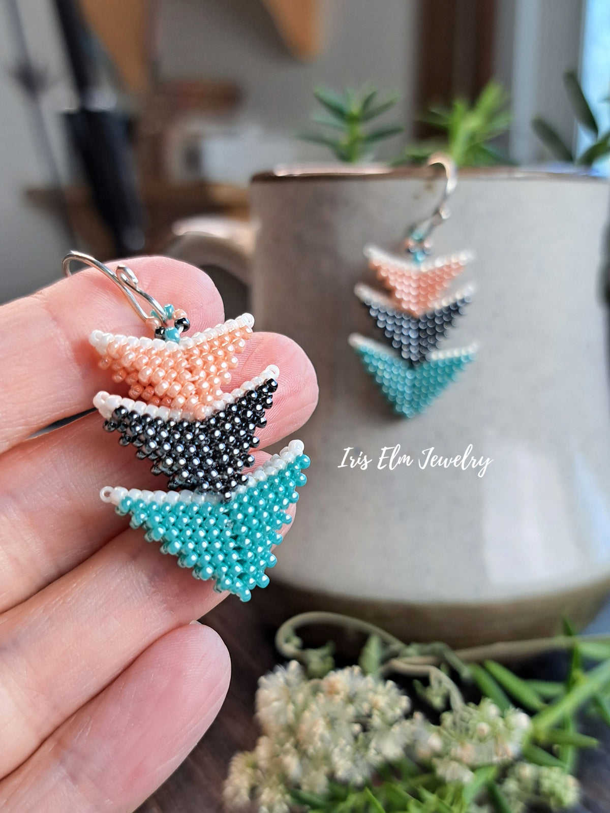 A hand holds a pair of handmade beaded earrings with a geometric chevron pattern in peach, grey, and turquoise, with a ceramic mug and dried flowers in the background.
