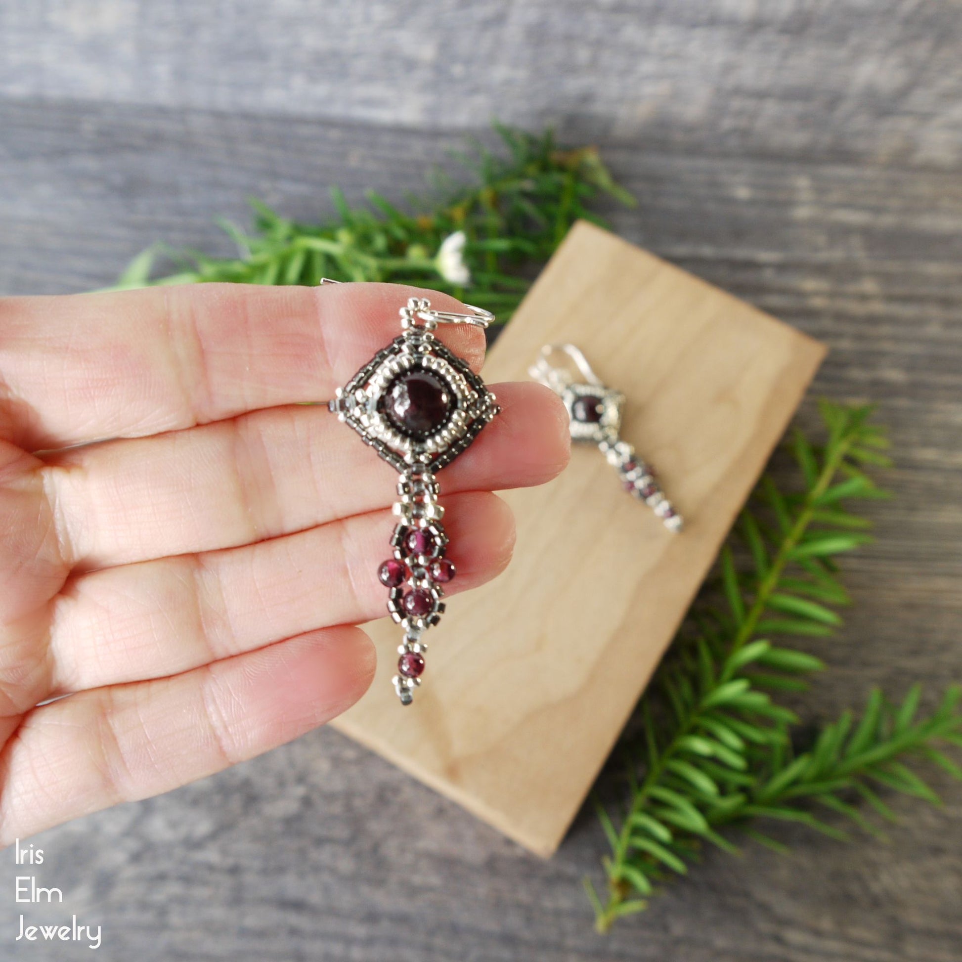 Hand holding a decorative silver and red garnet gemstone earring with sterling silver ear wires with a wooden box and greenery in the background.