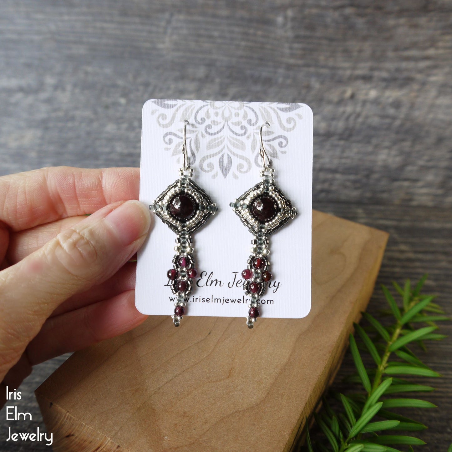 Pair of garnet beadwork earrings on a 'Iris Elm Jewelry' card held by a hand with a wooden surface and greenery in the background.