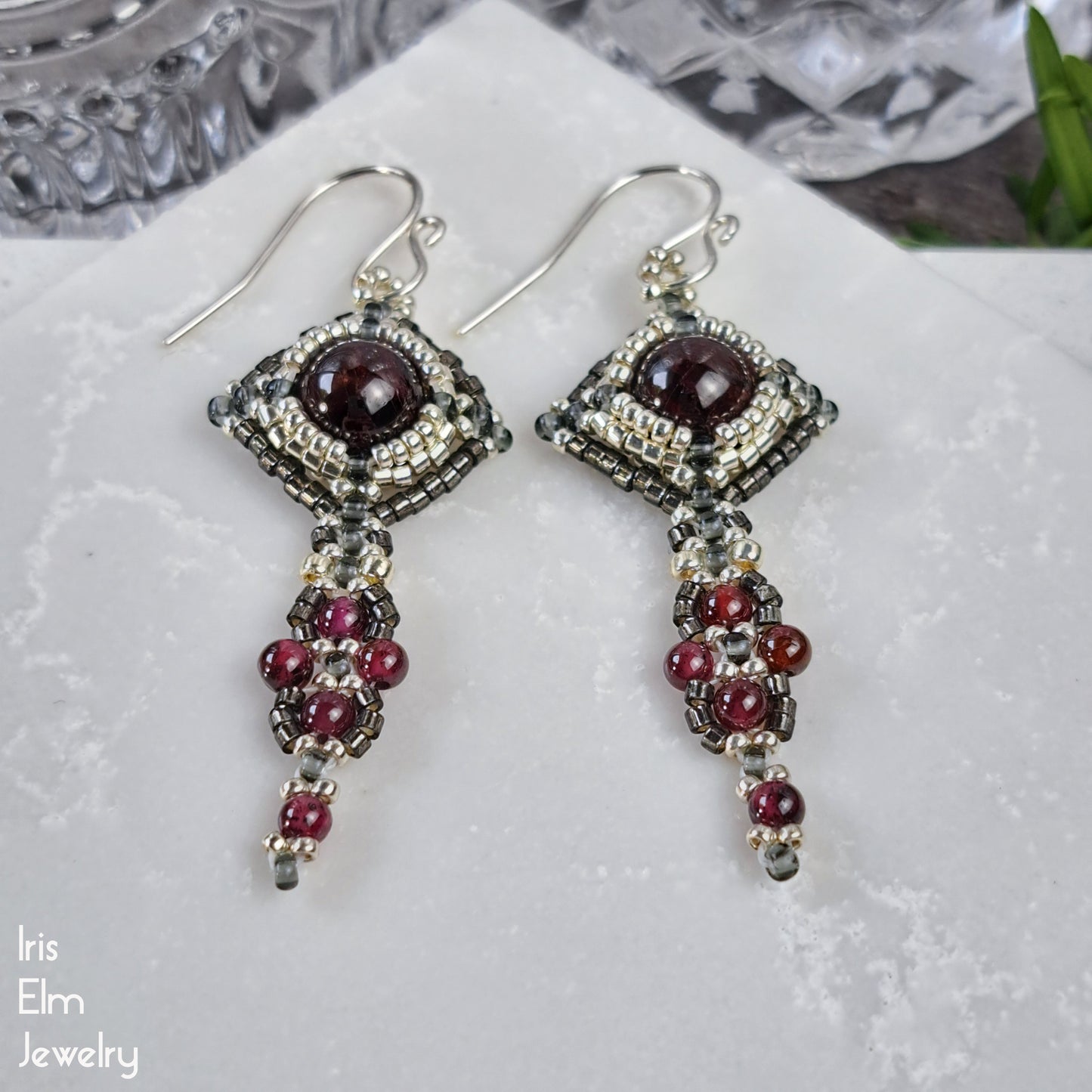 handmade beadwork Pair of silver art nouveau style earrings with red garnet gemstones on a white surface, branded 'Iris Elm Jewelry'.