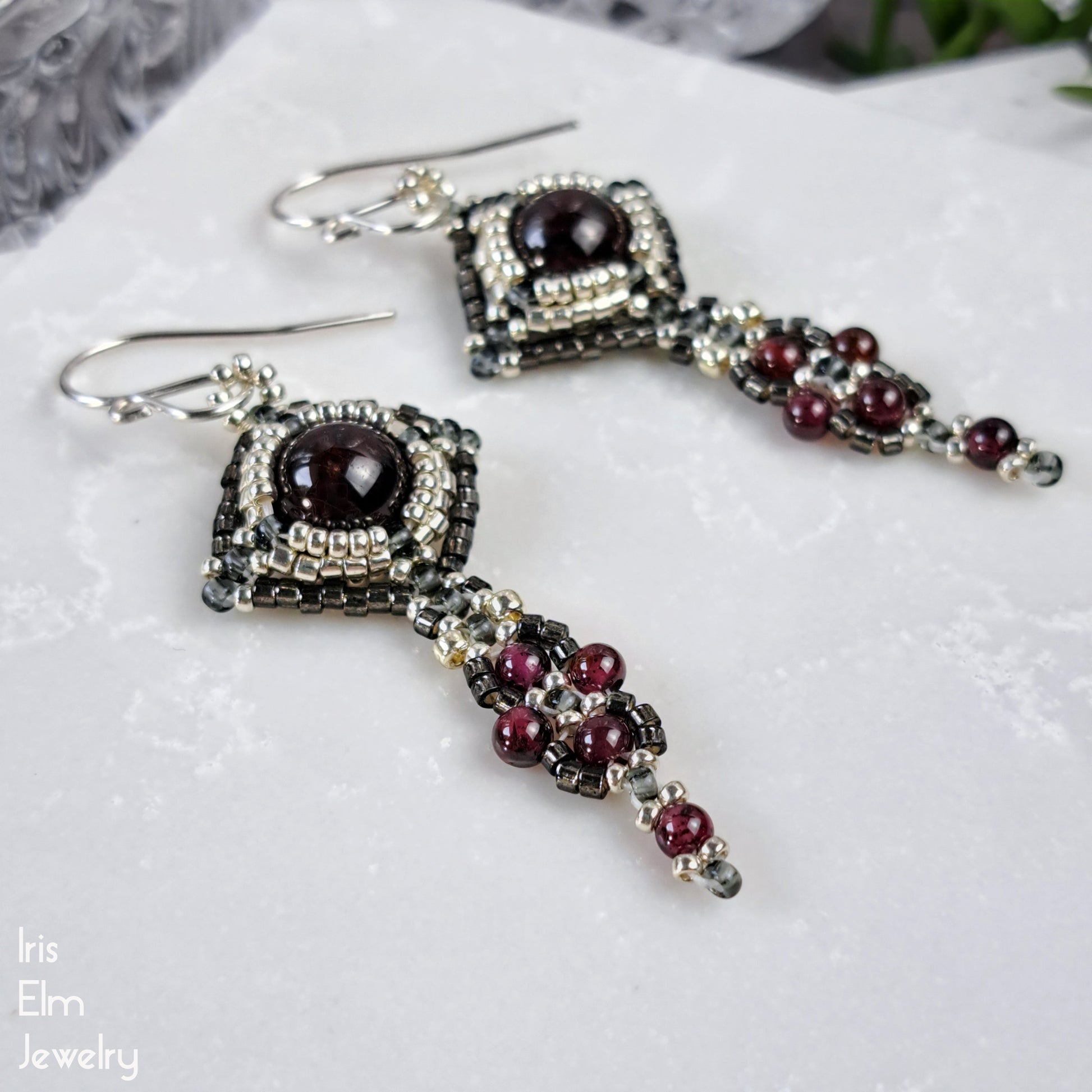 Ornate earrings with dark red garnet gemstones and glass bead accents on a white surface, branded 'Iris Elm Jewelry'.