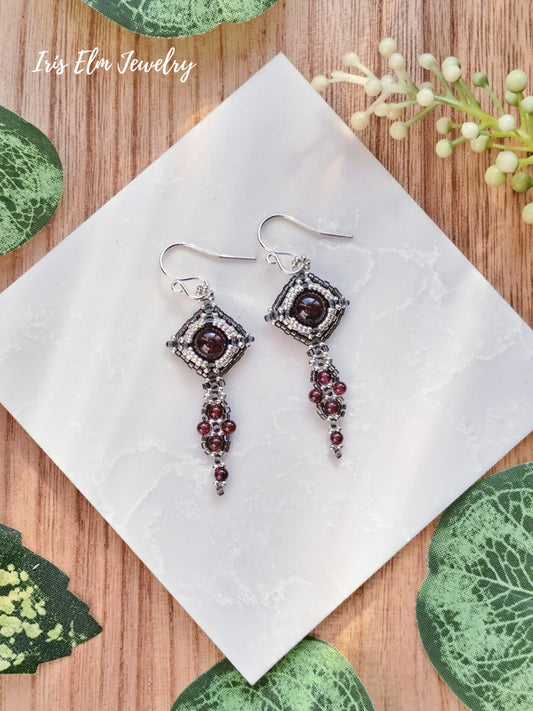 Pair of silver beadwork garnet art nouveau earrings with red gemstones on a white tile with green leaf patterns.