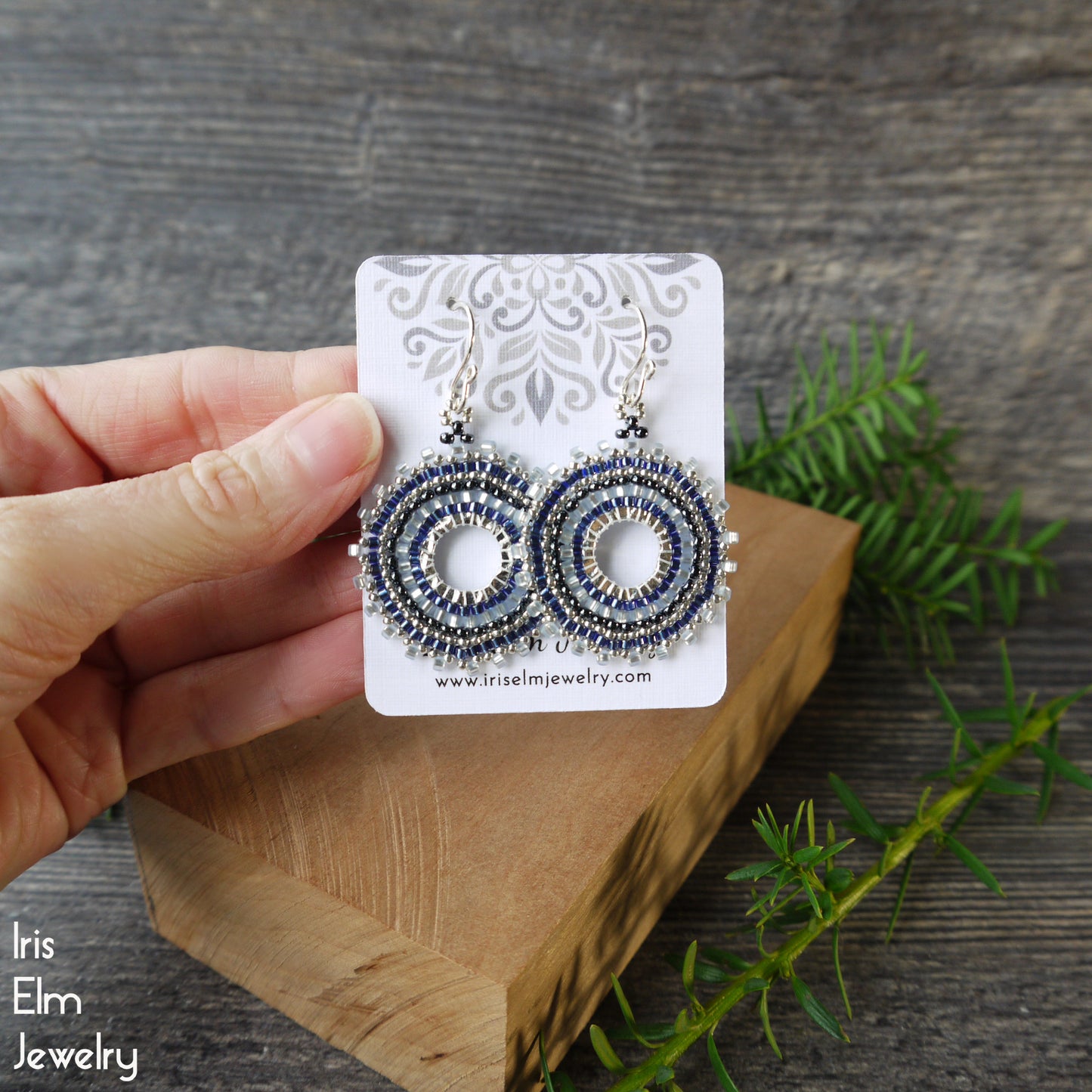 Hand holding a pair of beaded woven blue hoop earrings on a branded card with a wooden block and greenery in the background.