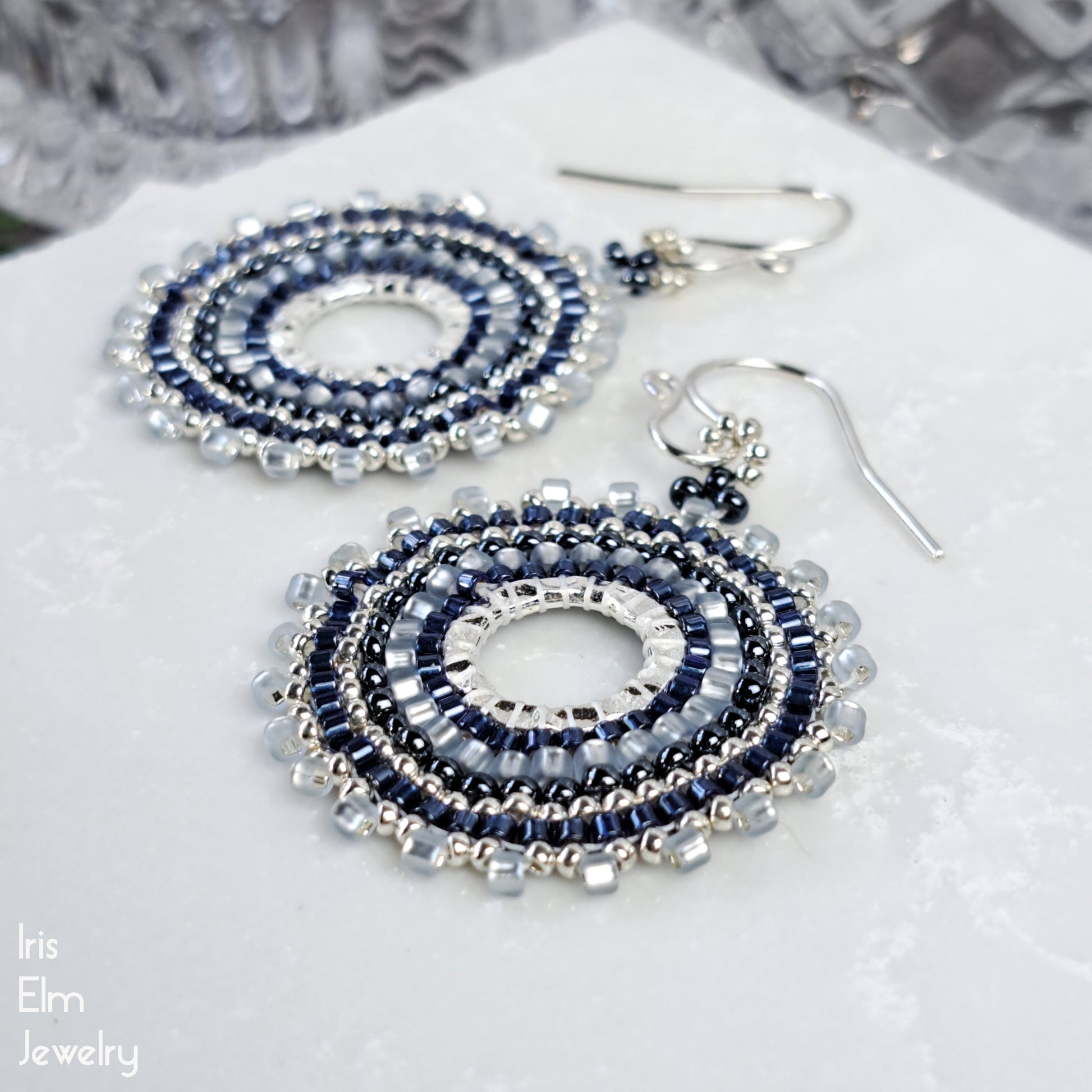 Beaded seed bead hoop handcrafted blue earrings on a white surface with 'Iris Elm Jewelry' branding.