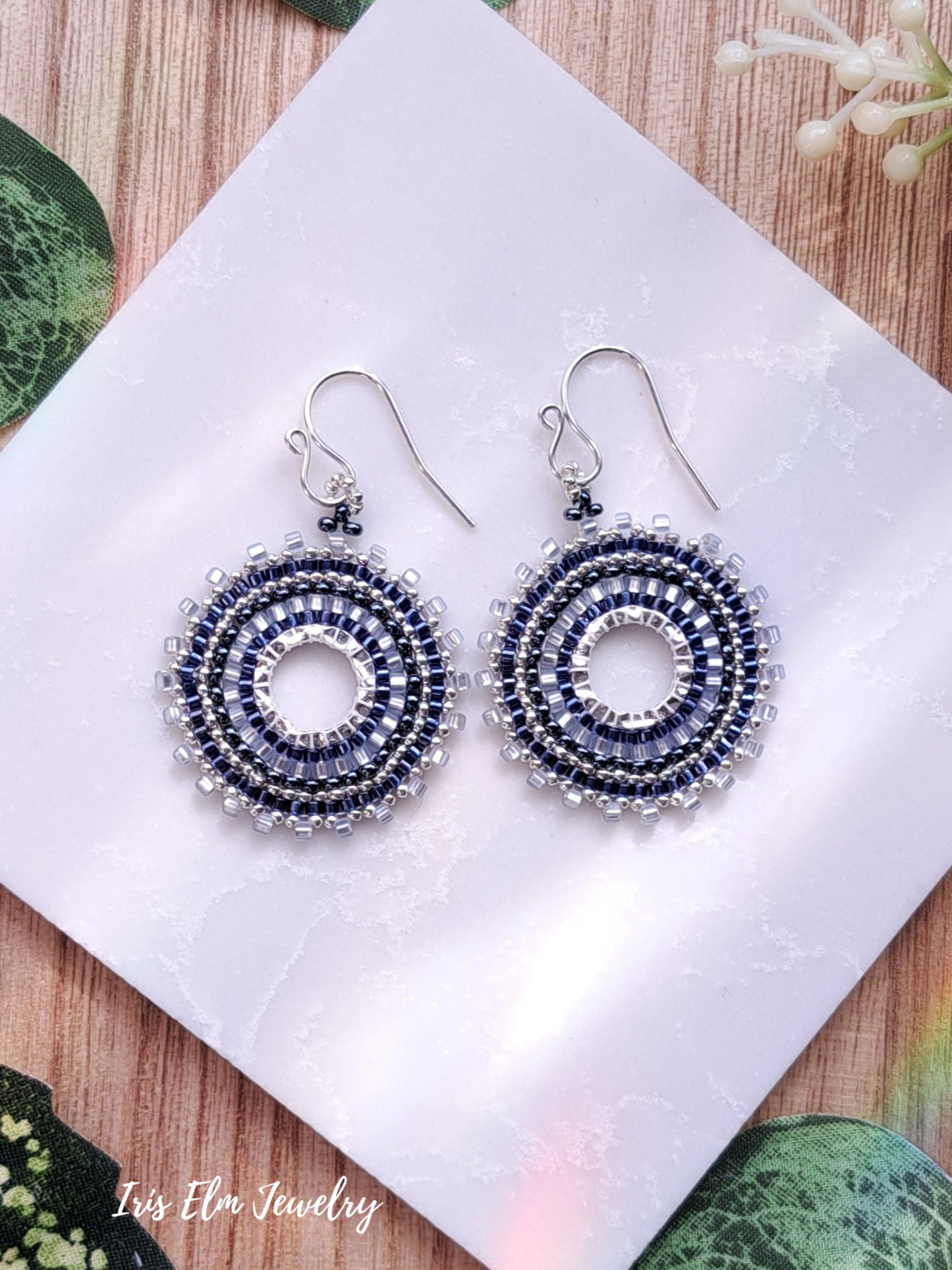 Pair of beaded blue seed bead woven hoop earrings on a white cloth with 'Iris Elm Jewelry' branding.