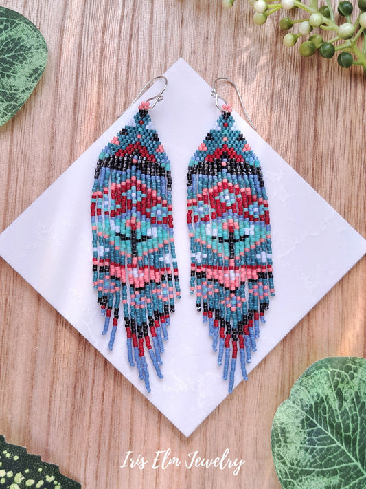 Colorful beaded floral folk art blue red coral mint seed bead fringe earrings on a white card with green leaves around, featuring 'Iris Elm Jewelry'.