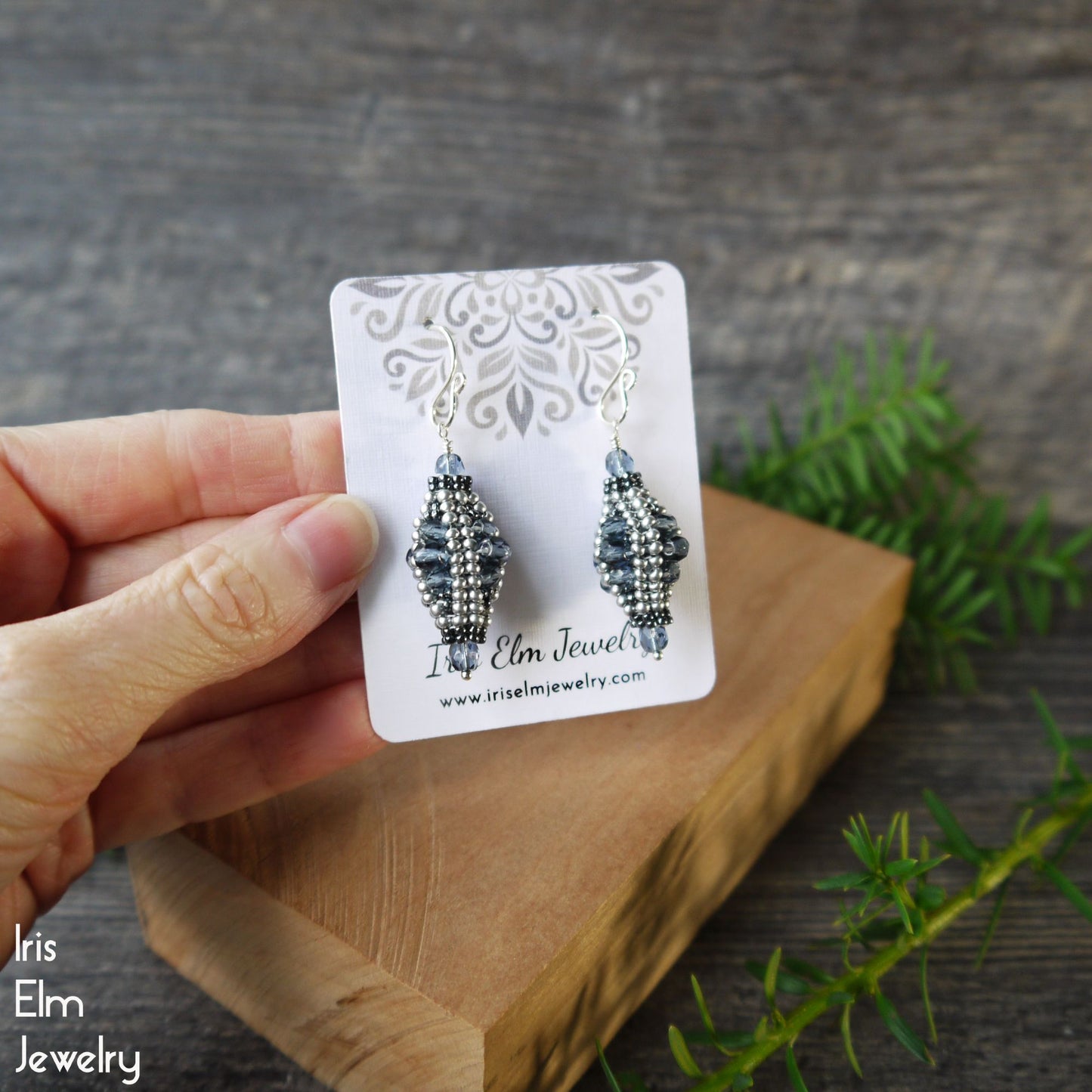 Pair of beaded blue glass drop earrings on a 'Iris Elm Jewelry' card held by a hand with a wooden block and greenery in the background.
