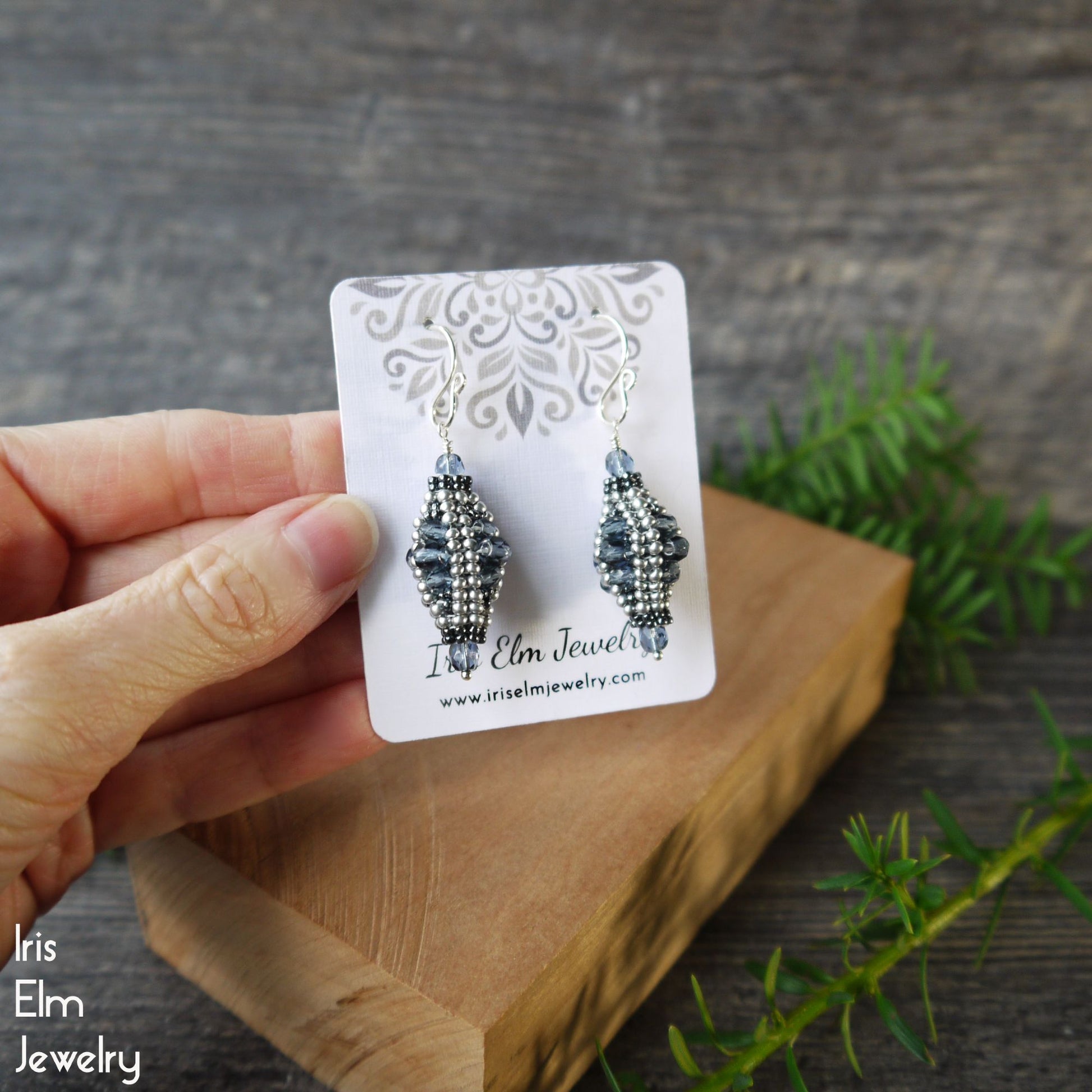 Pair of beaded blue glass drop earrings on a 'Iris Elm Jewelry' card held by a hand with a wooden block and greenery in the background.