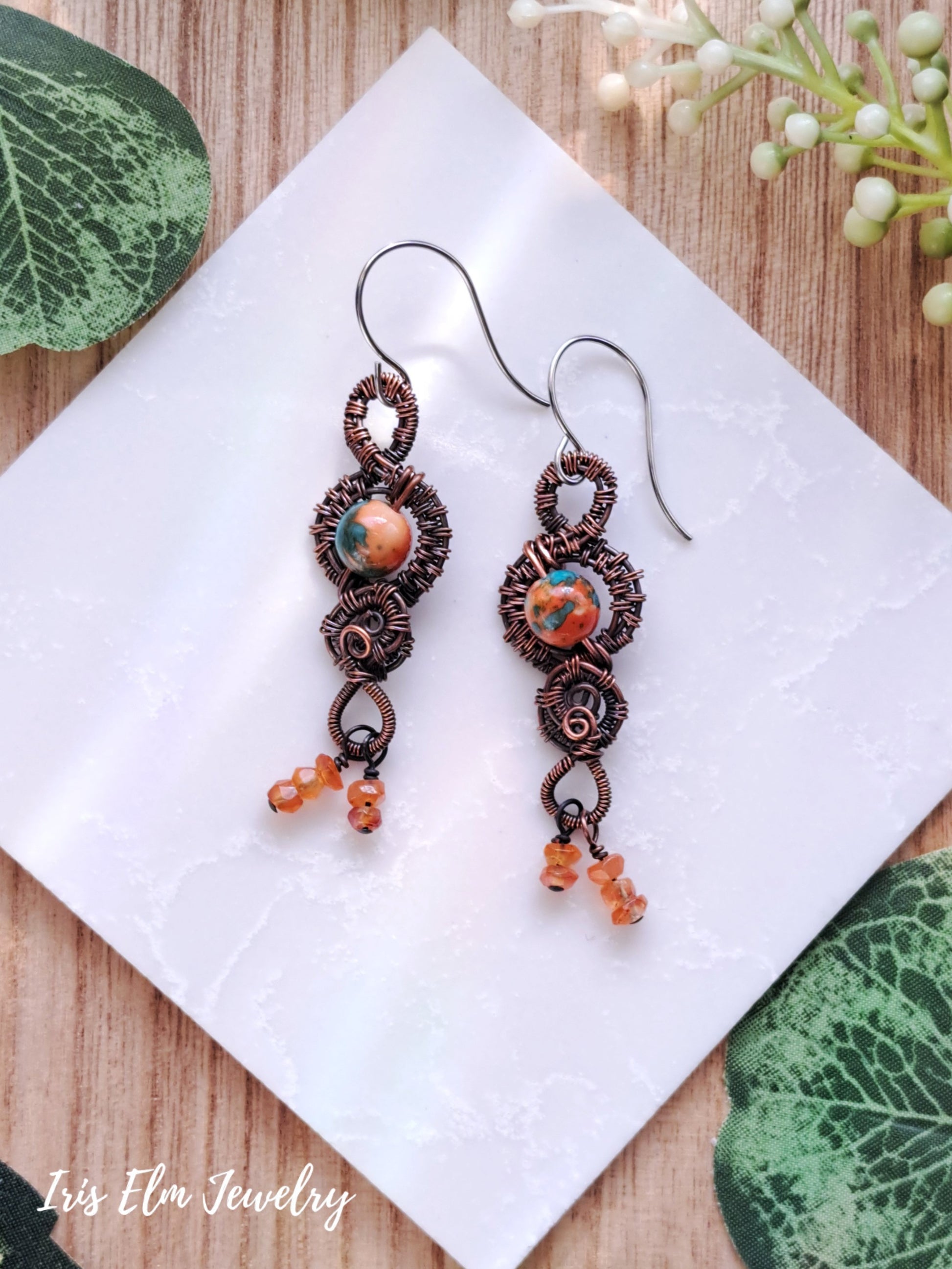 Pair of copper wire-wrapped orange ocean jade and carnelian earrings with orange beads on a white tile.