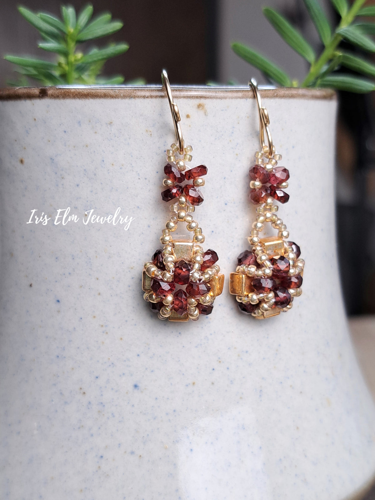 a pair of handmade red garnet gemstone and gold glass beaded earrings by Iris Elm Jewelry dangling from ceramic pottery