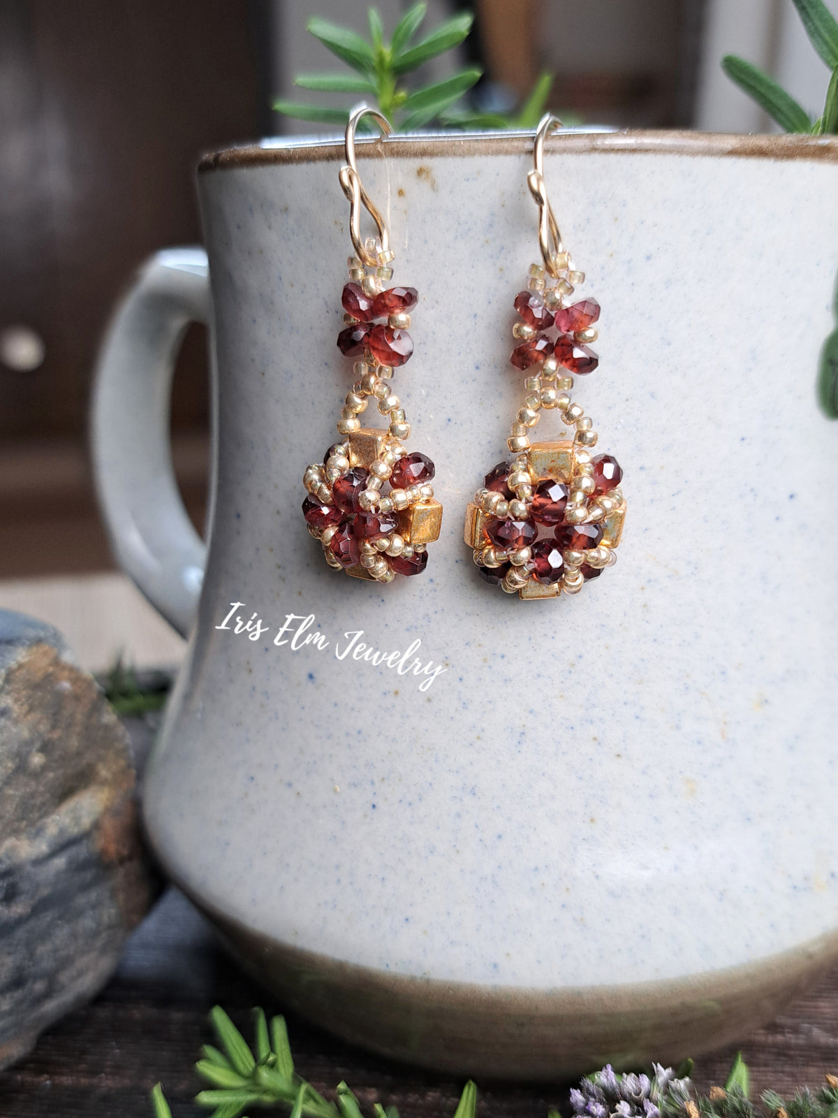 a pair of handmade red garnet gemstone and gold glass beaded earrings by Iris Elm Jewelry dangling from ceramic pottery
