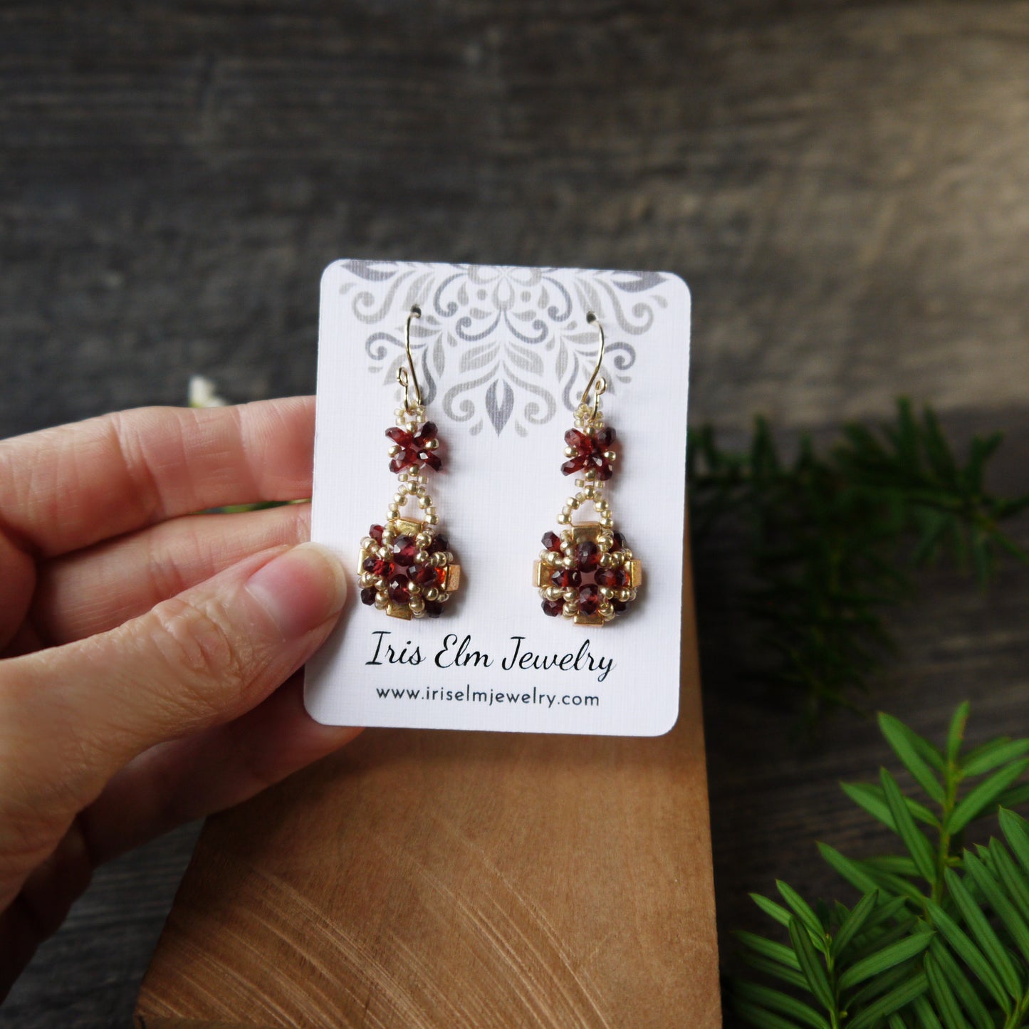 Pair of beaded garnet earrings on a 'Iris Elm Jewelry' card held by a hand with a wooden surface and greenery in the background.