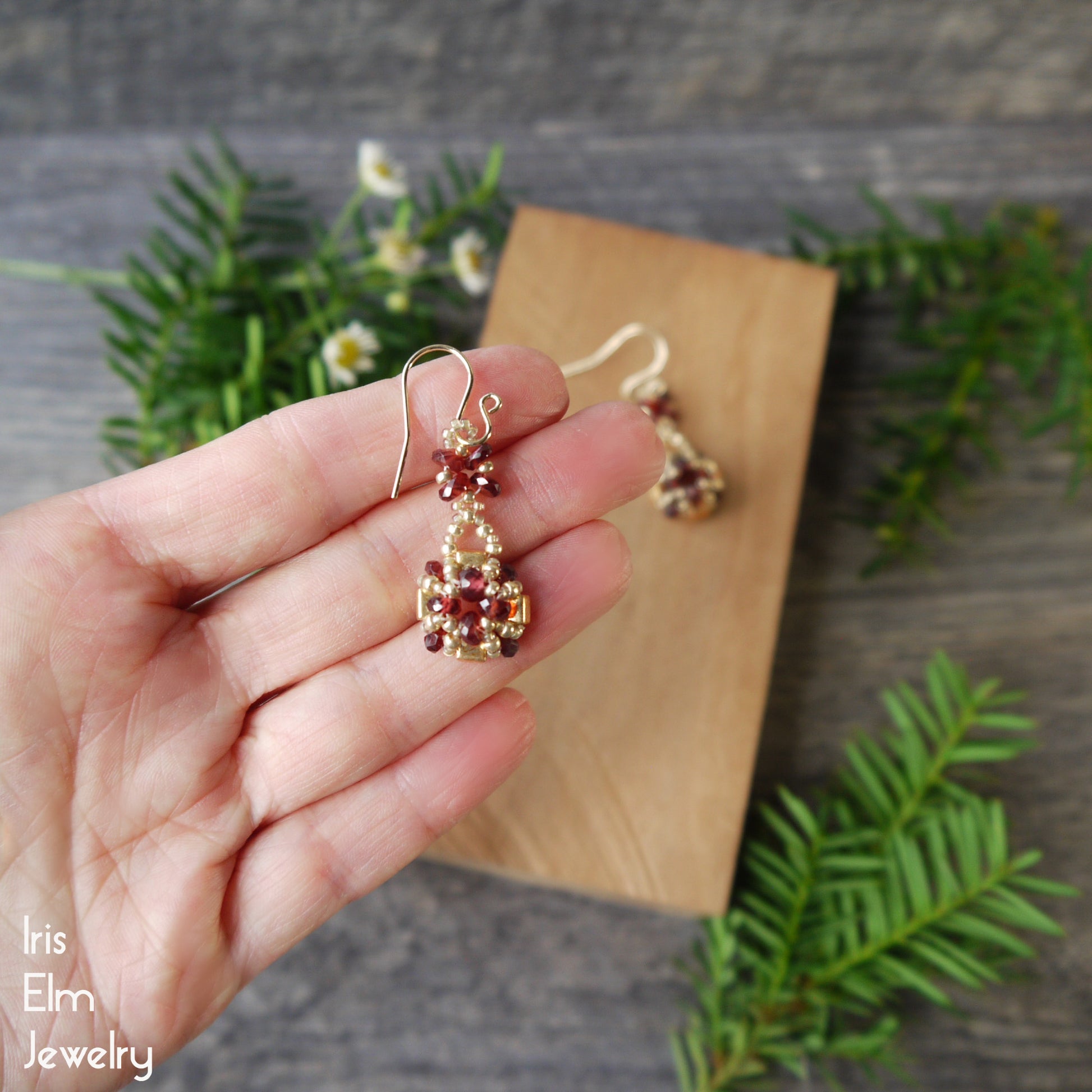 Gold and red garnet beaded earrings held in a hand with a wooden block and greenery in the background.