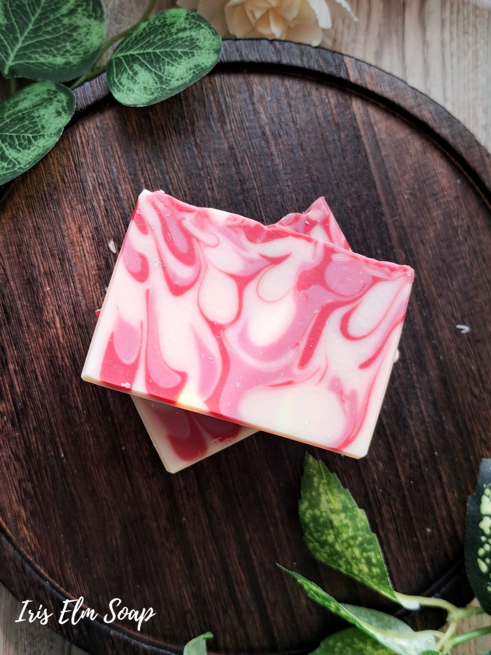 Two pink and white marbled strawberry peach handmade soap bars on a wooden surface with green leaves around.