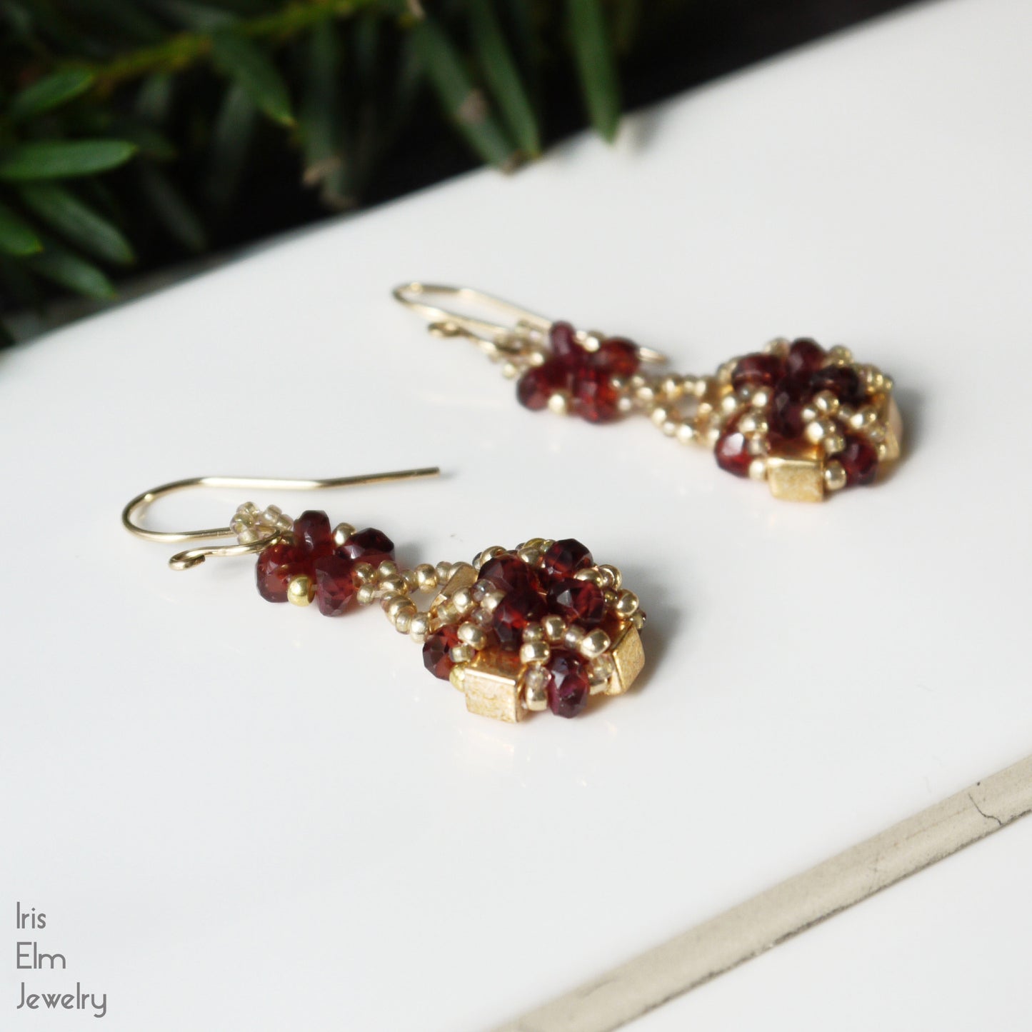 Pair of gold and red garnet gemstone beaded earrings on a white surface with greenery in the background.