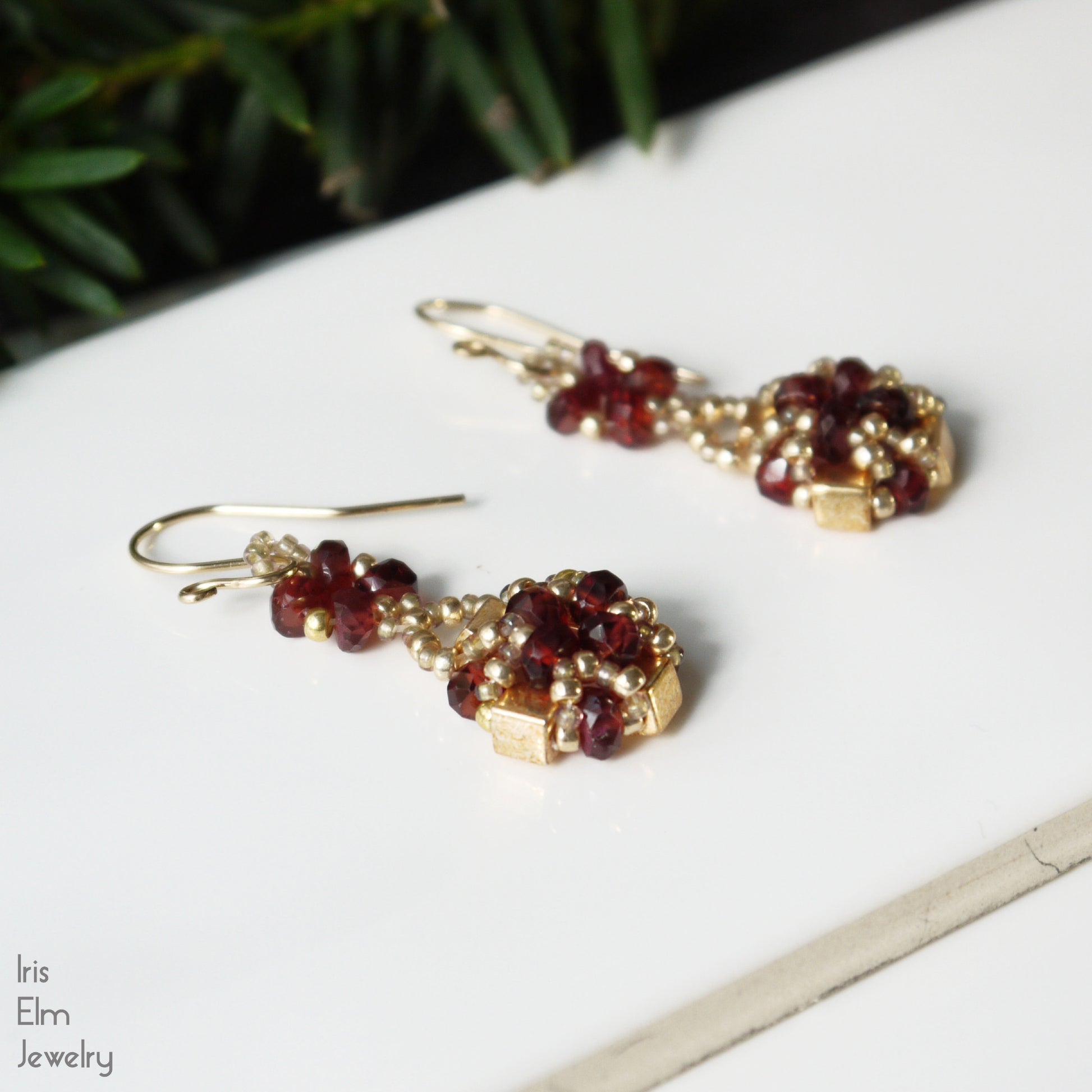 Pair of gold and red garnet gemstone beaded earrings on a white surface with greenery in the background.