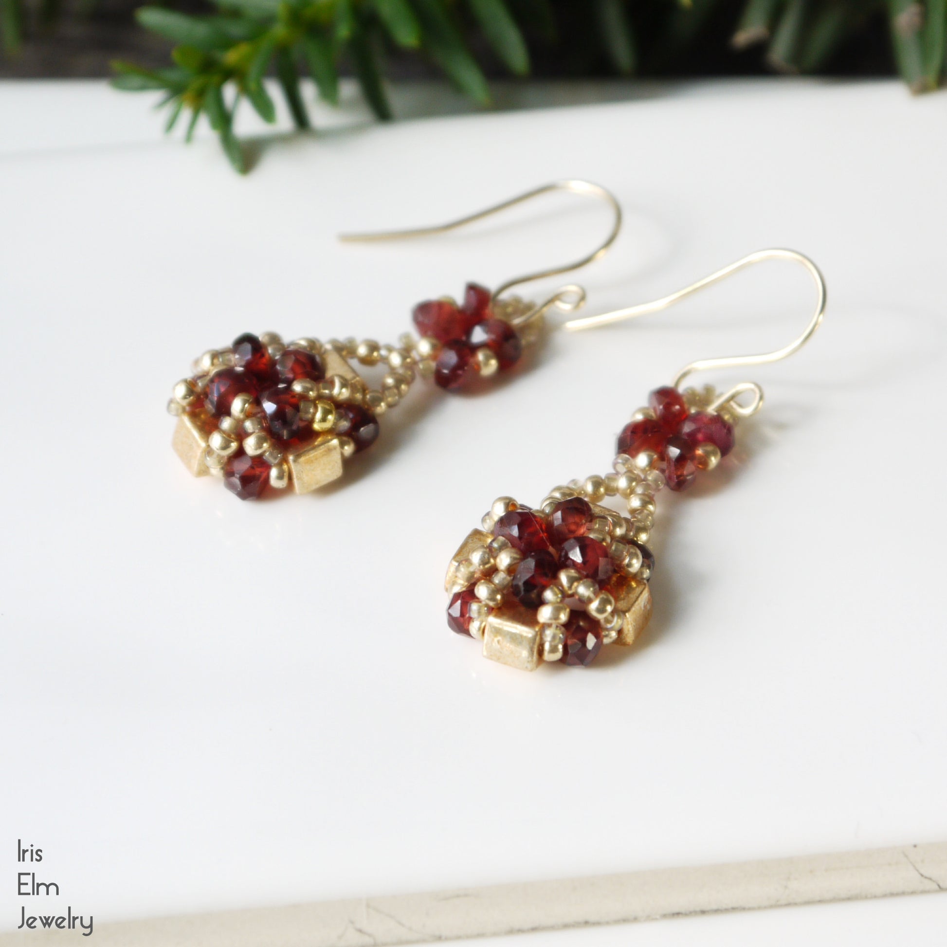 Pair of gold glass and red garnet stone beaded earrings on a white surface with greenery in the background.