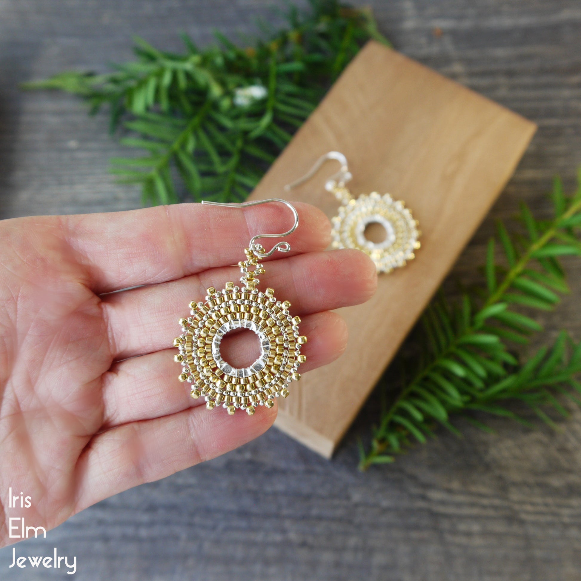 Gold beaded seed bead hoop earrings held in a hand with a wooden block and greenery in the background, featuring 'Iris Elm Jewelry' branding.