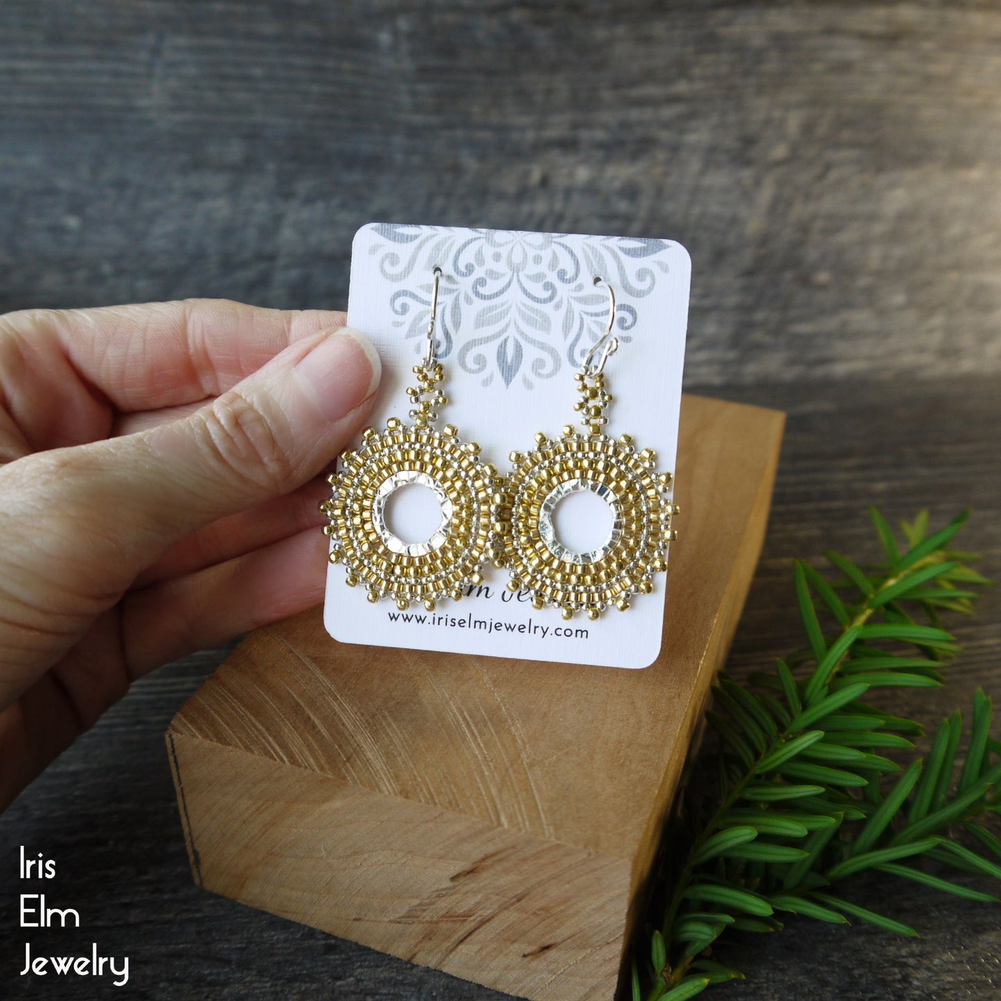 Gold hoop earrings on a card held by a hand with a wooden block and greenery in the background