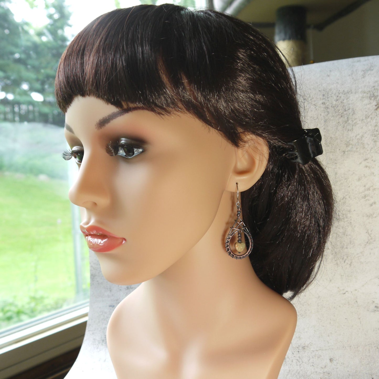 Mannequin head with a black wig and copper teardrop earrings on a neutral background