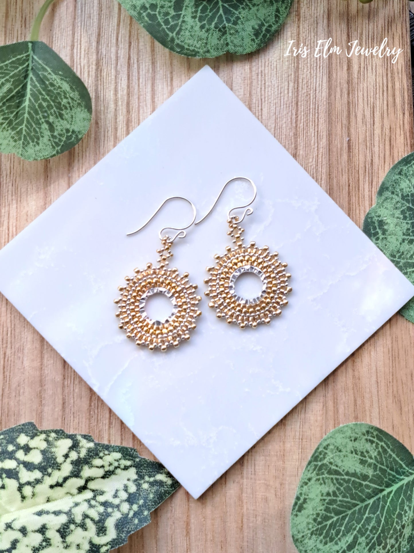 Gold circular seed bead woven hoop earrings on a white card with green leaves around
