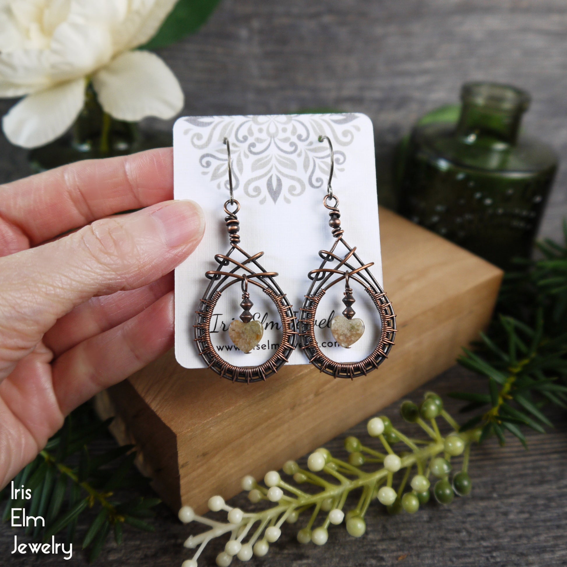 copper wire woven teardrop earrings with heart shaped autumn jasper stones in center on a card held by a hand with a decorative background