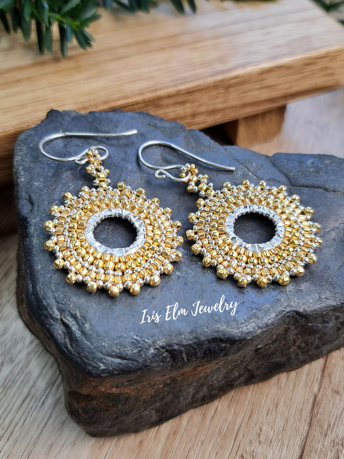 A pair of handmade beaded dangle earrings with gold and silver elements, displayed on a dark stone slab with a wooden background. The brand name 'Iris Elm Jewelry' is printed on the stone.