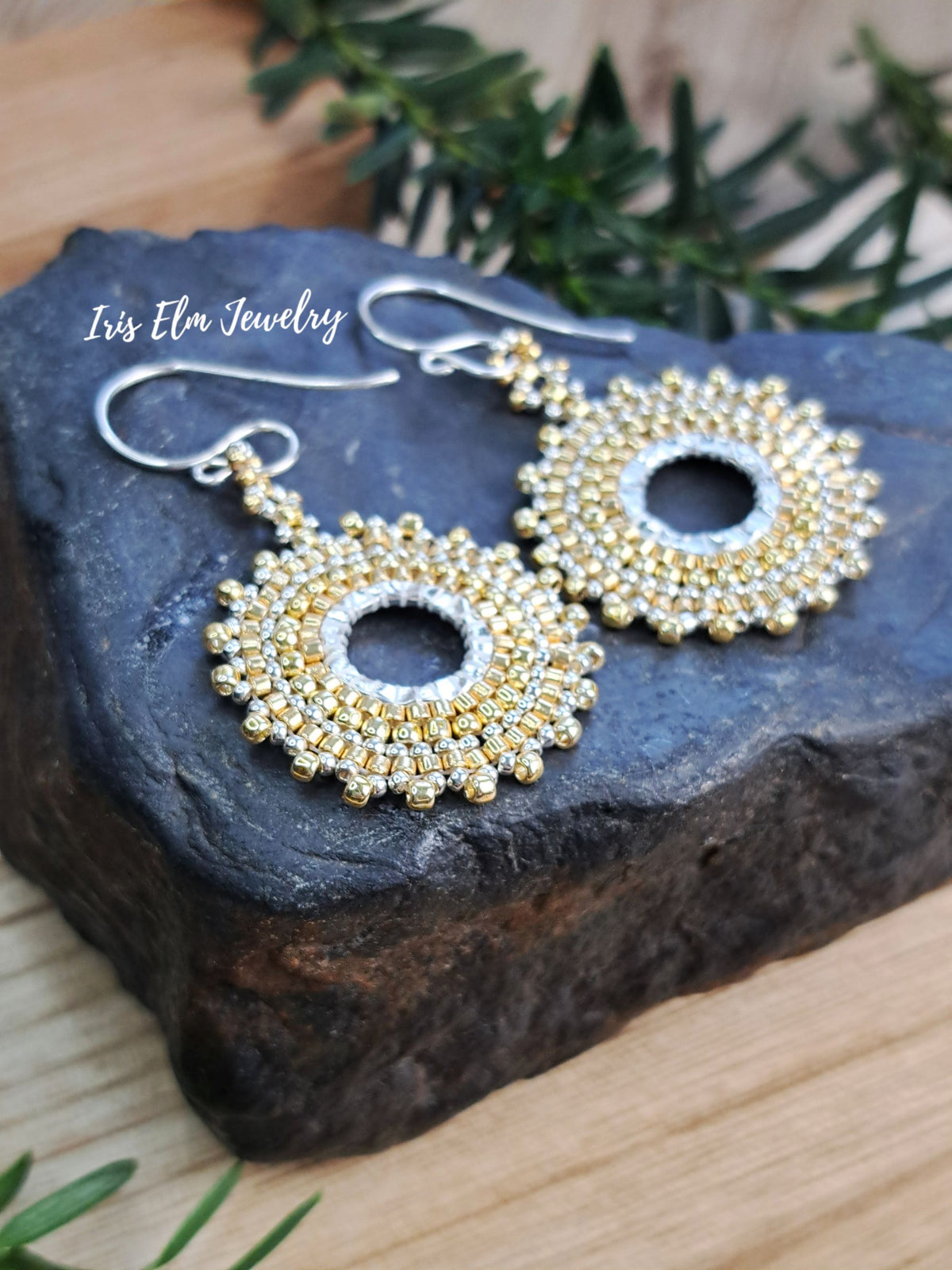 A pair of gold and silver beaded dangle earrings displayed on a dark stone slab with green foliage in the background. The brand name 'Iris Elm Jewelry' is printed in white text on the upper left.