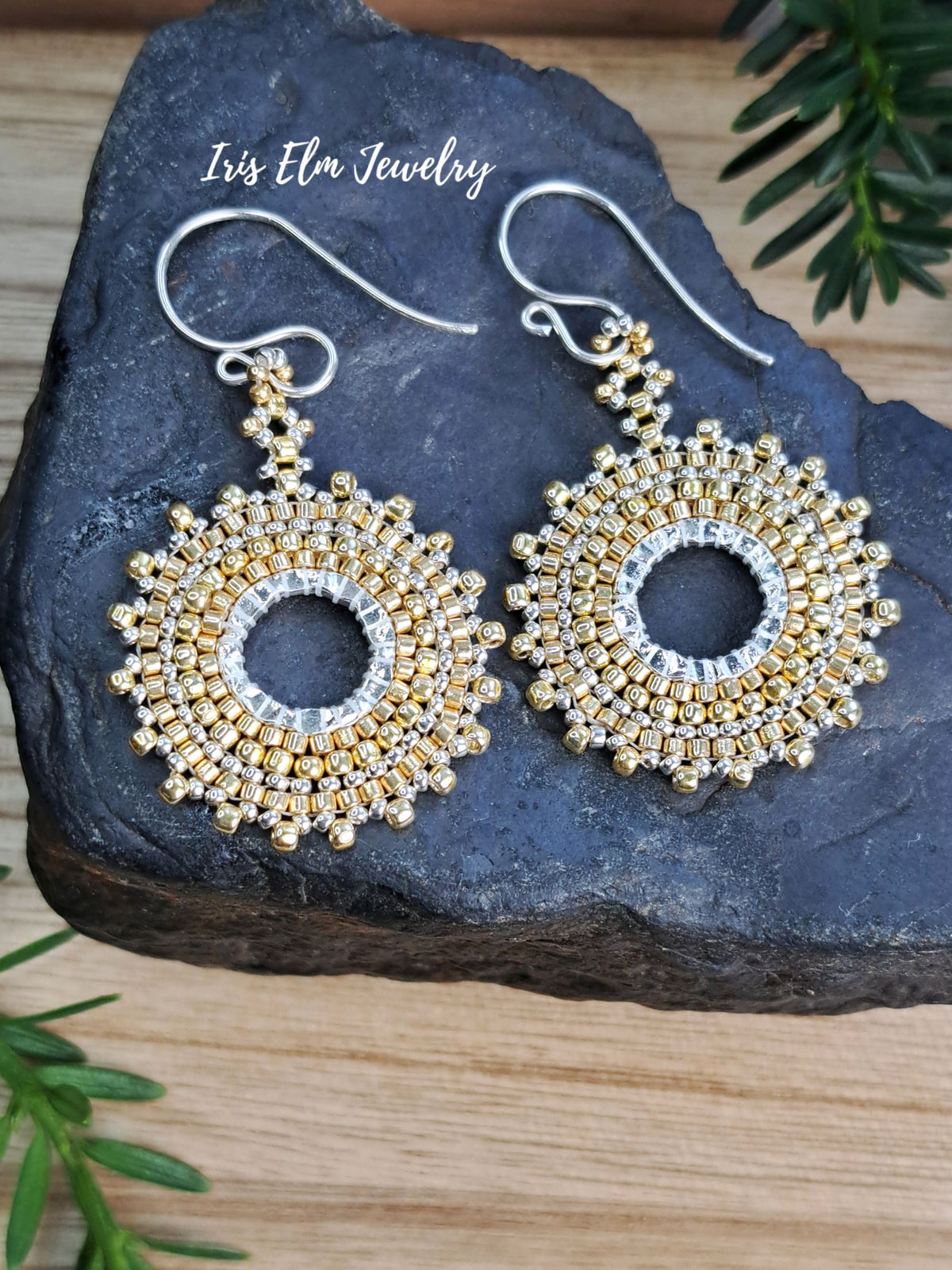 A pair of beaded dangle earrings displayed on a dark stone slab over a wooden surface, with green pine branches in the corners. The earrings feature intricate gold and silver seed bead circular sunburst designs with silver fishhook ear wires.