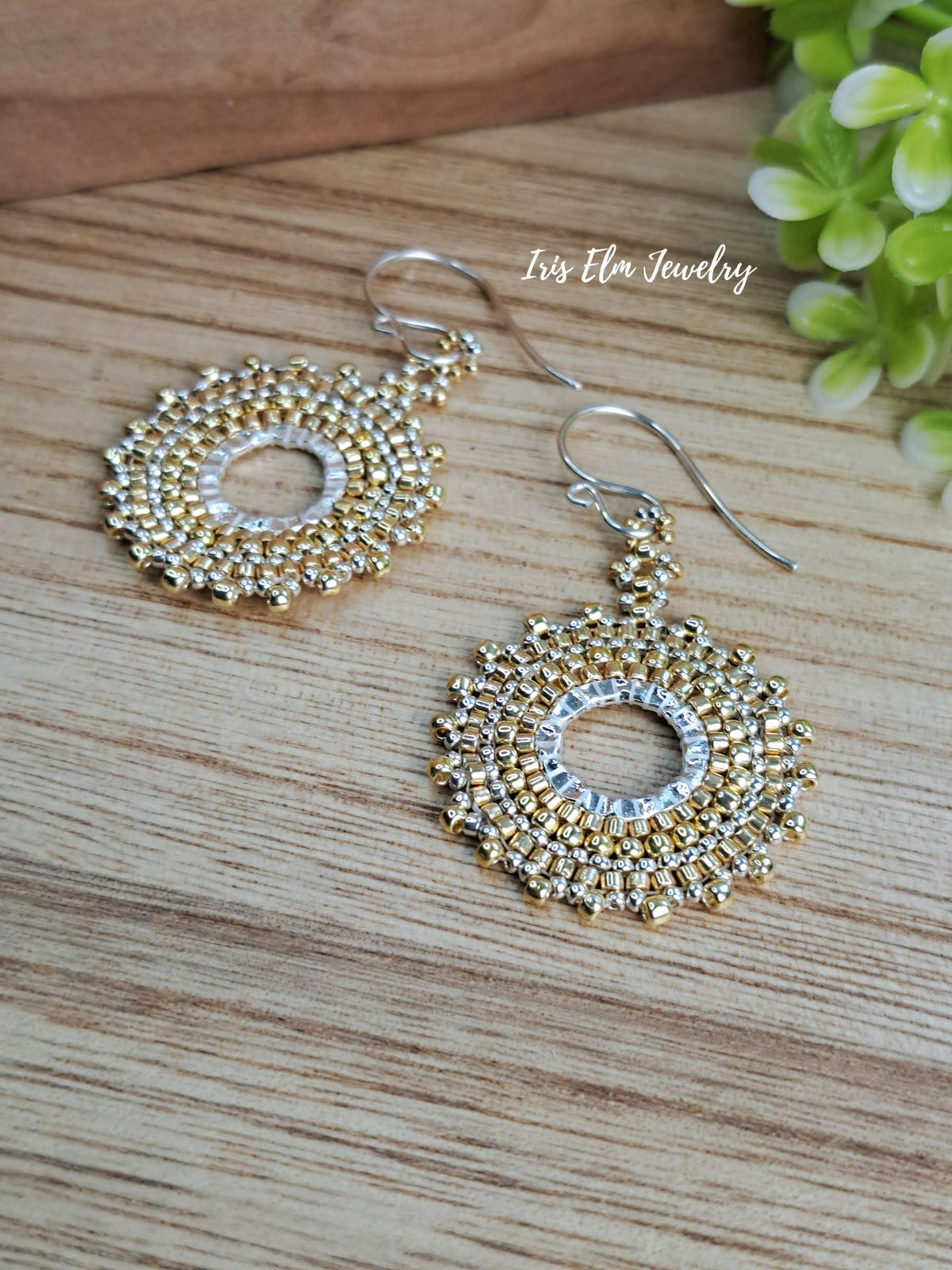A pair of handmade beaded dangle earrings with gold and silver metallic beads, displayed on a light wood surface with a green plant in the background. The brand name 'Iris Elm Jewelry' is watermarked on the image.