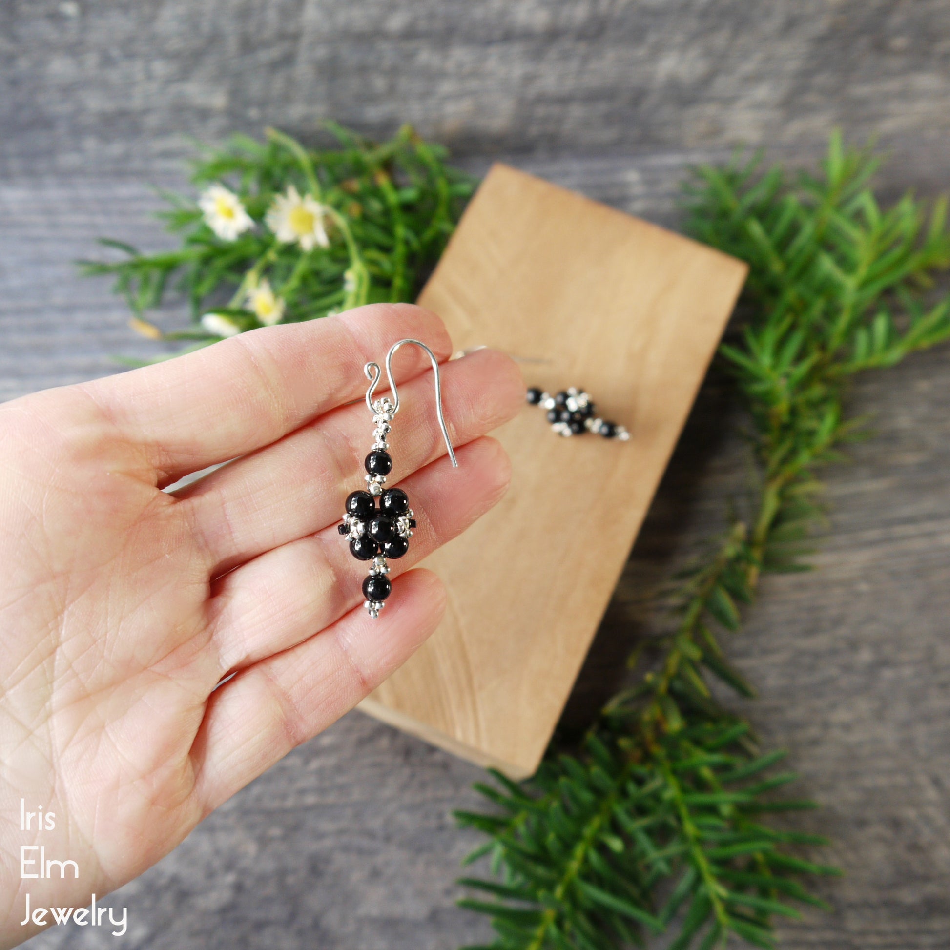 Hand holding a pair of black beaded onyx stone earrings with sterling silver ear wires with a wooden box and greenery in the background.