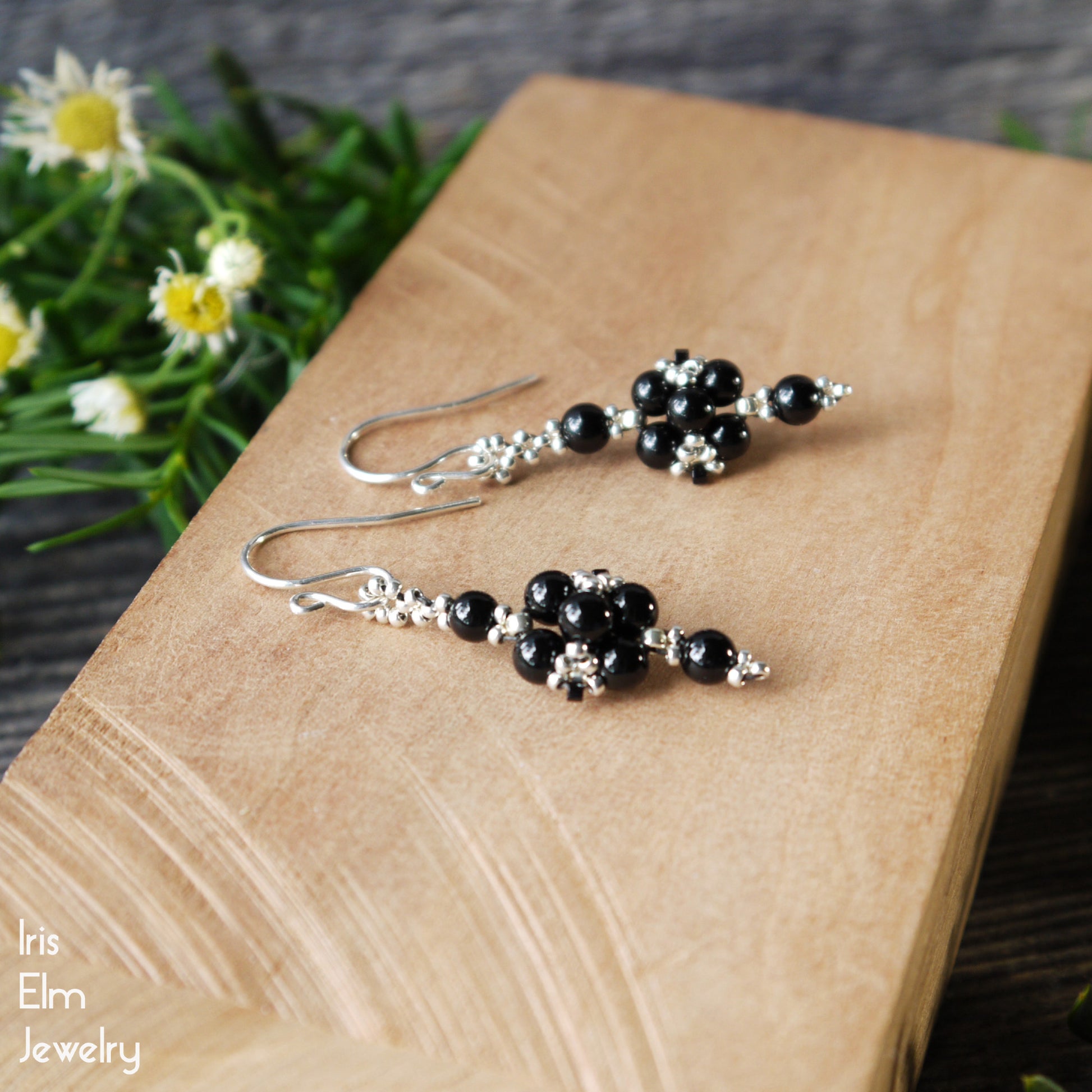 handmade Black onyx beaded earrings on a wooden surface with flowers in the background, featuring 'Iris Elm Jewelry' branding.