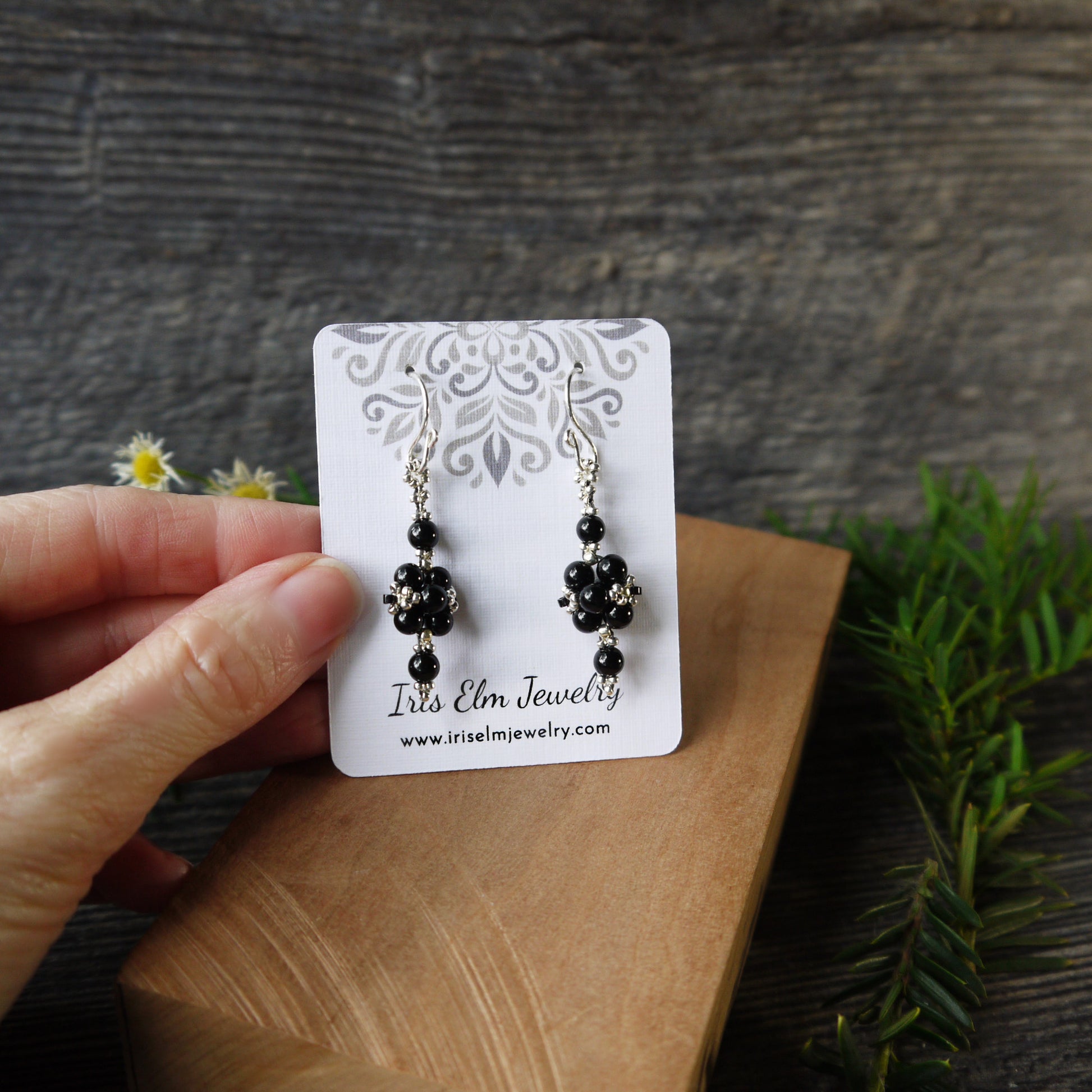 Black onyx and silver beaded earrings on a 'Iris Elm Jewelry' card held by a hand with a wooden surface and plants in the background.