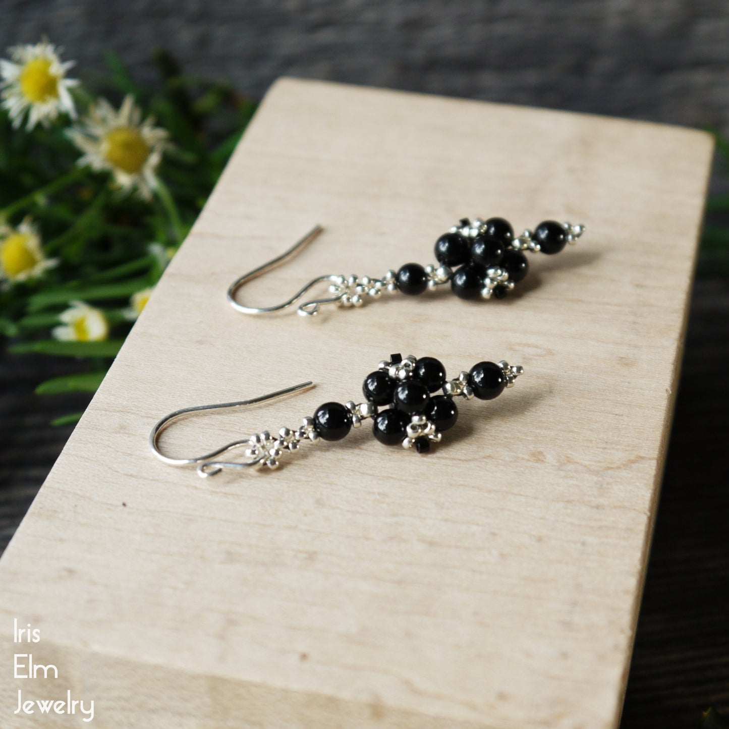 Black beaded stone handcrafted beadwork earrings on a wooden surface with flowers in the background, branded 'Iris Elm Jewelry'.