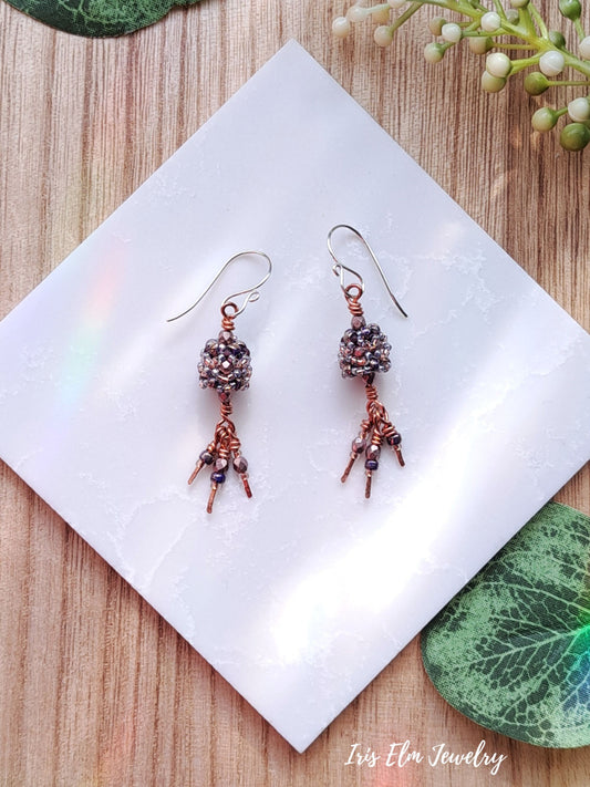 Pair of light purple copper dangle earrings on a white square with green leaves in the background