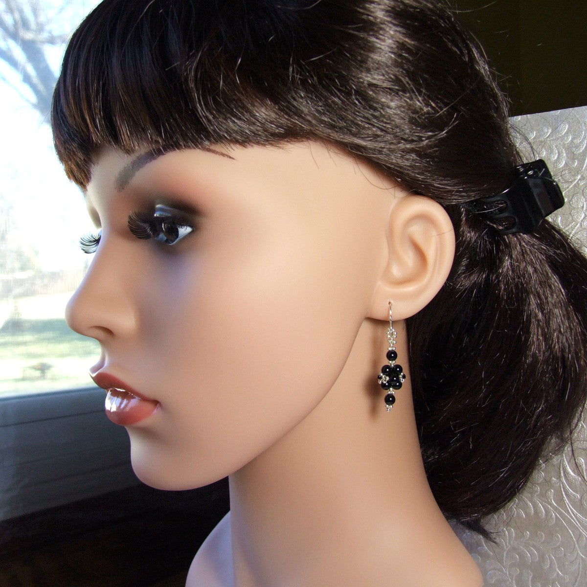 Mannequin head with black hair and small black onyx earrings, set against a blurred outdoor background.